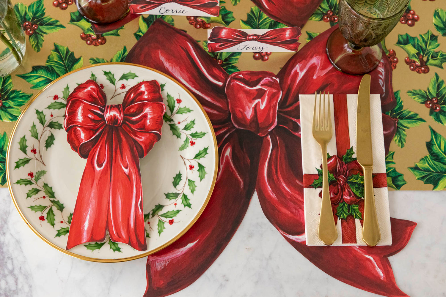 An elegant table setting with the Gilded Holly Runner underneath the Die-Cut Bow Placemat, gold silverware on the Christmas Bow Guest Napkin and Christmas Bow Table accent on a holly dinner plate.