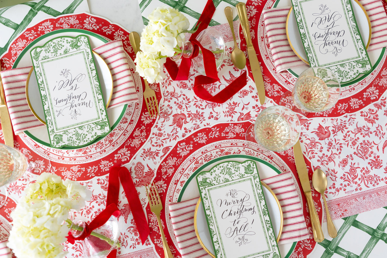 A table set for three featuring the Die-cut Red Regal Placemat, green rimmed plates with Green Regal Peacock Table Accents atop, and gold flatware on the Red Regal Peacock Runner.