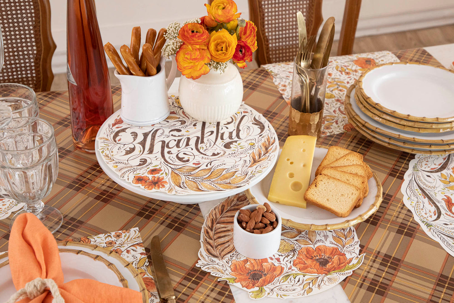 A fall-themed dining table with a &quot;Thankful&quot; serving paper, floral centerpiece, plates of cheese and bread, almonds, breadsticks, and cutlery. The autumn plaid-patterned tablecloth ties together these Fall Essentials beautifully.