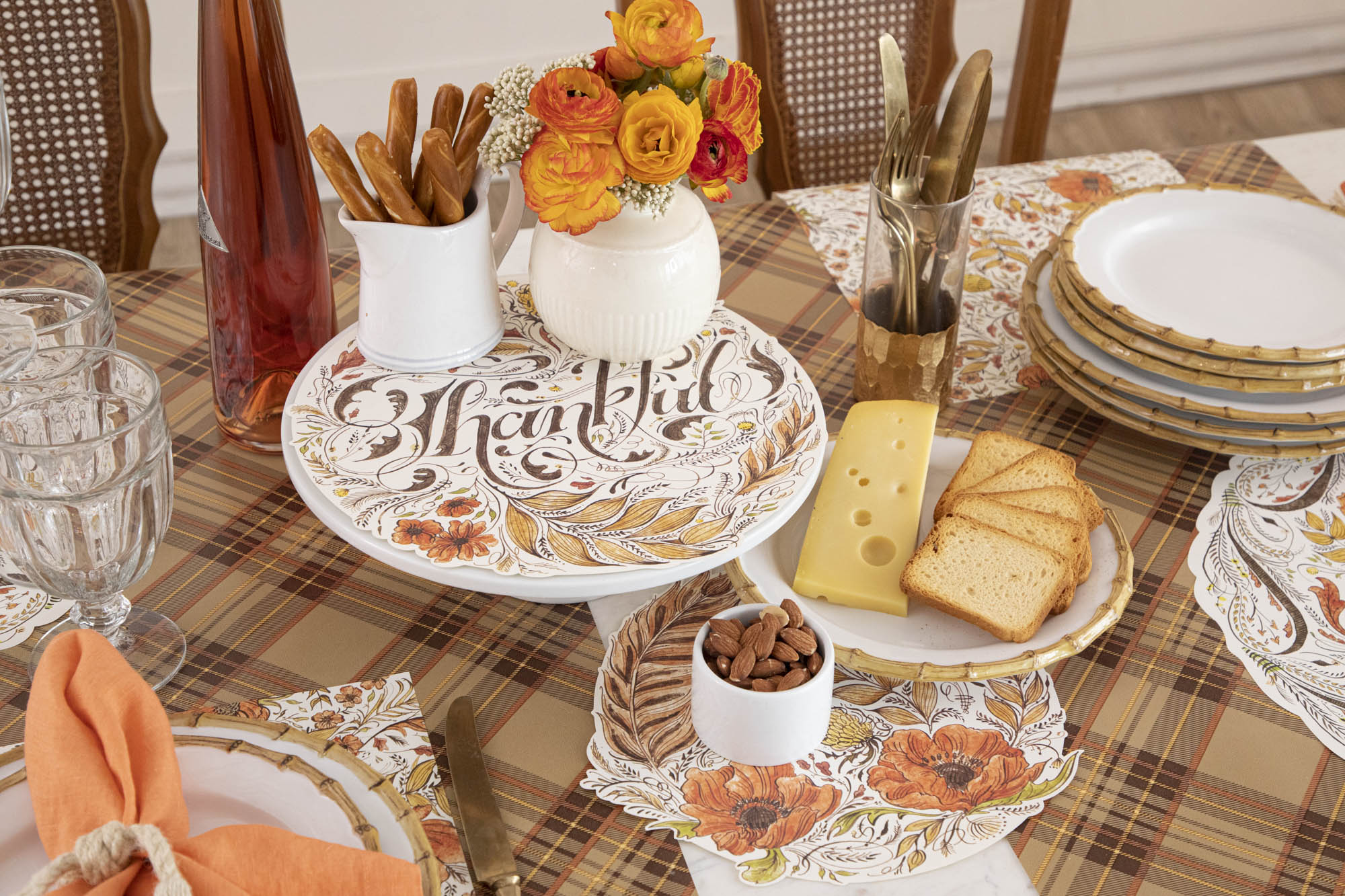 A fall-themed dining table with a "Thankful" serving paper, floral centerpiece, plates of cheese and bread, almonds, breadsticks, and cutlery. The autumn plaid-patterned tablecloth ties together these Fall Essentials beautifully.