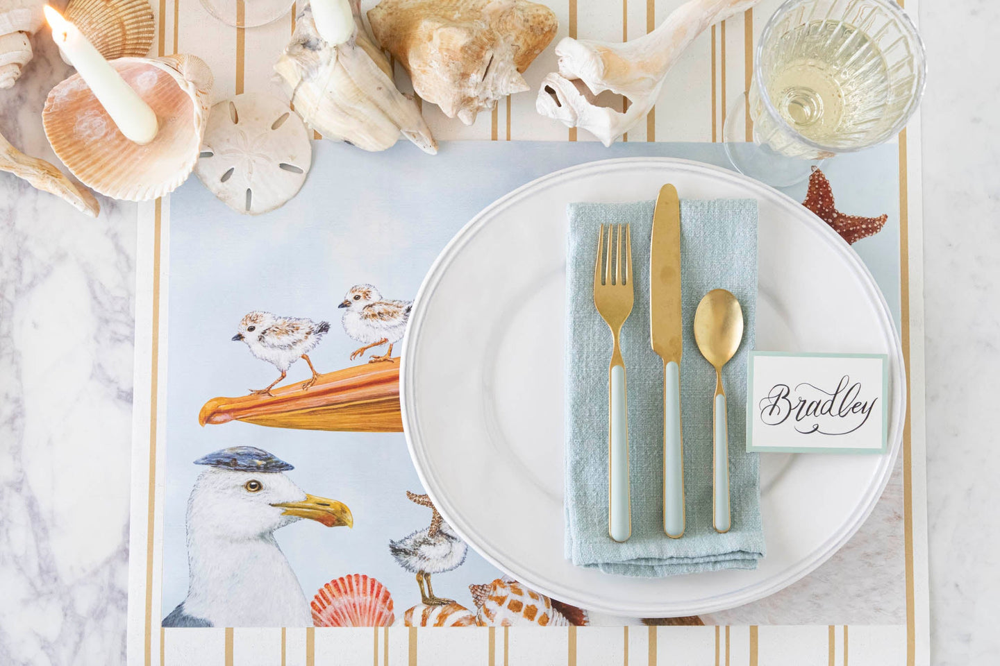A sea-themed table setting with the Shorebirds Placemat, seafoam colors, Seafoam Frame Place Card and seashells on the Antique Gold Stripe Runner.