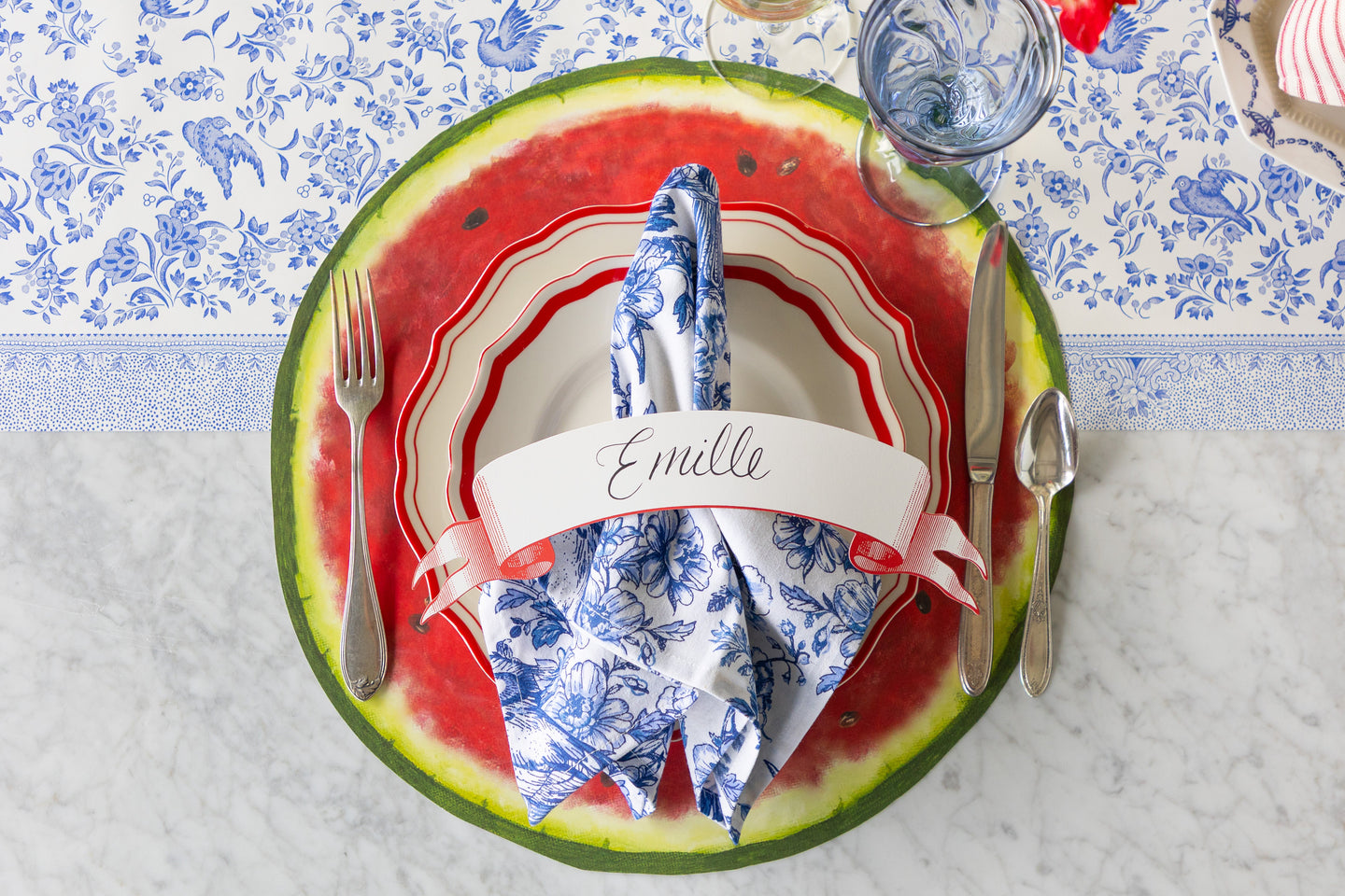 A place setting featuring the Die-cut Watermelon Placemat, red scalloped plates with the Bluebird Floral Napkin and Classic Red Banner Table Accent atop, and vintage silverware on the Blue Regal Peacock Runner.