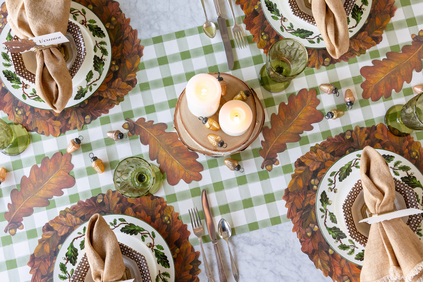 Moss Painted Check Runner under a fall table setting with Die-cut Autumn Wreath Placemats, scattered Autumn Leaf Table Accents, acorn Ornaments and candles atop wood slices.