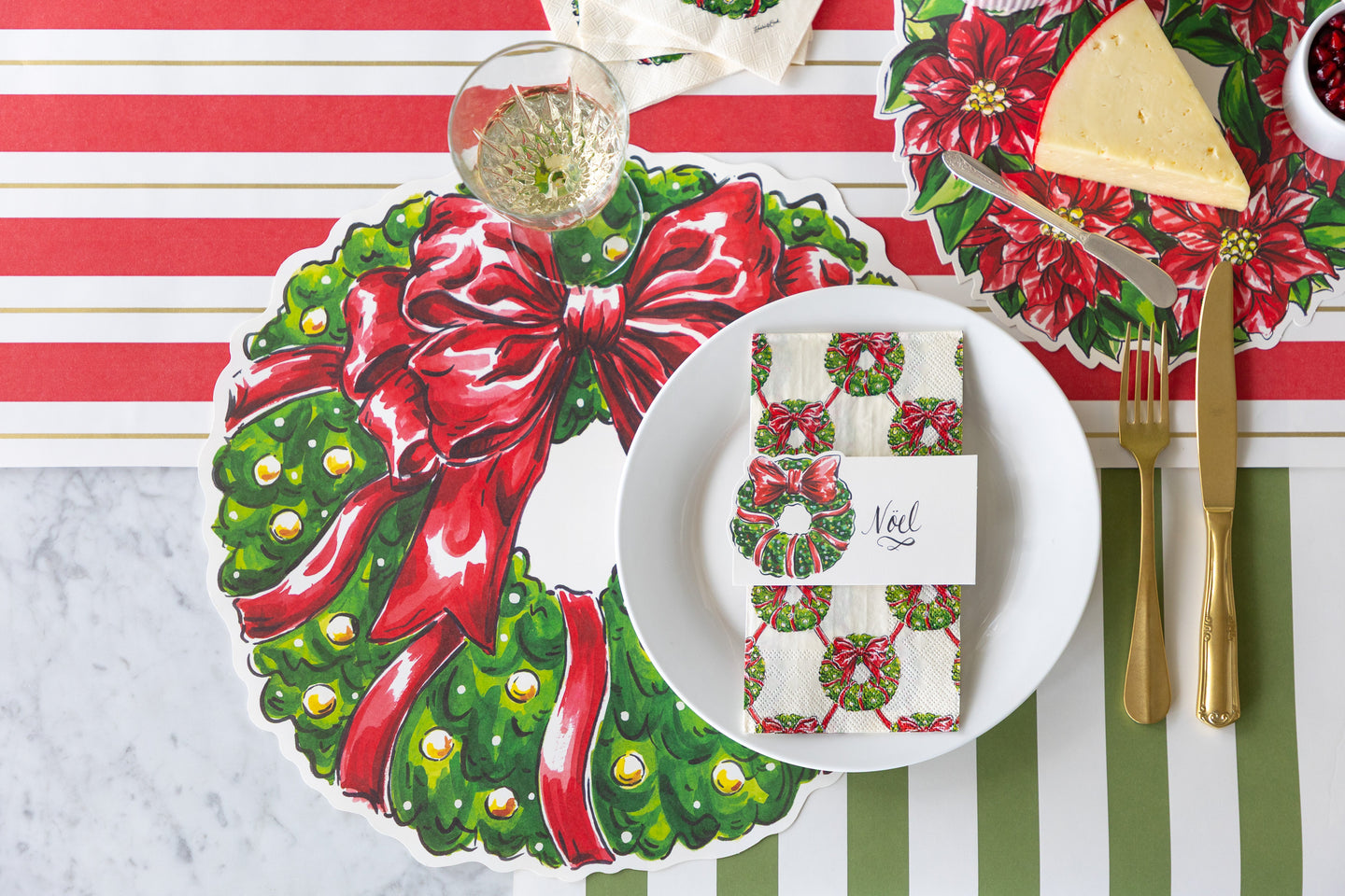 A place setting featuring the Die-cut Holiday Wreath Placemat, a plate with a Holiday Wreath Guest Napkin and Holiday Wreath Place Card atop, Holiday Wreath Serving Paper with a block of cheese, and Red & Gold Awning Stripe Runner underneath.