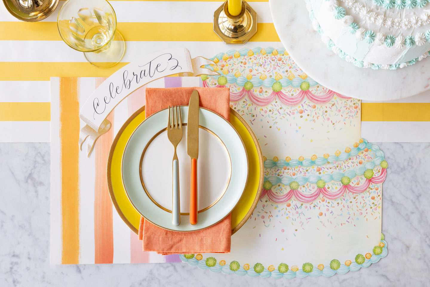 A birthday themed table setting featuring the Die-cut Birthday Cake Placemat, Sorbet Painted Stripe Placemat, colorful dinnerware, Classic Gold Banner Table Accent, decorative cake on the Marigold Stripe Runner.