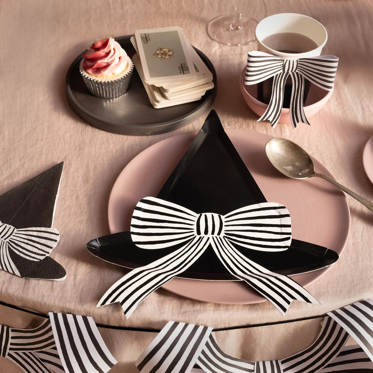 A table setting with black witch hat plates decorated with striped bows, a pink plate, a cupcake, black paper cups, playing cards, and striped bow decorations on a pink tablecloth.
