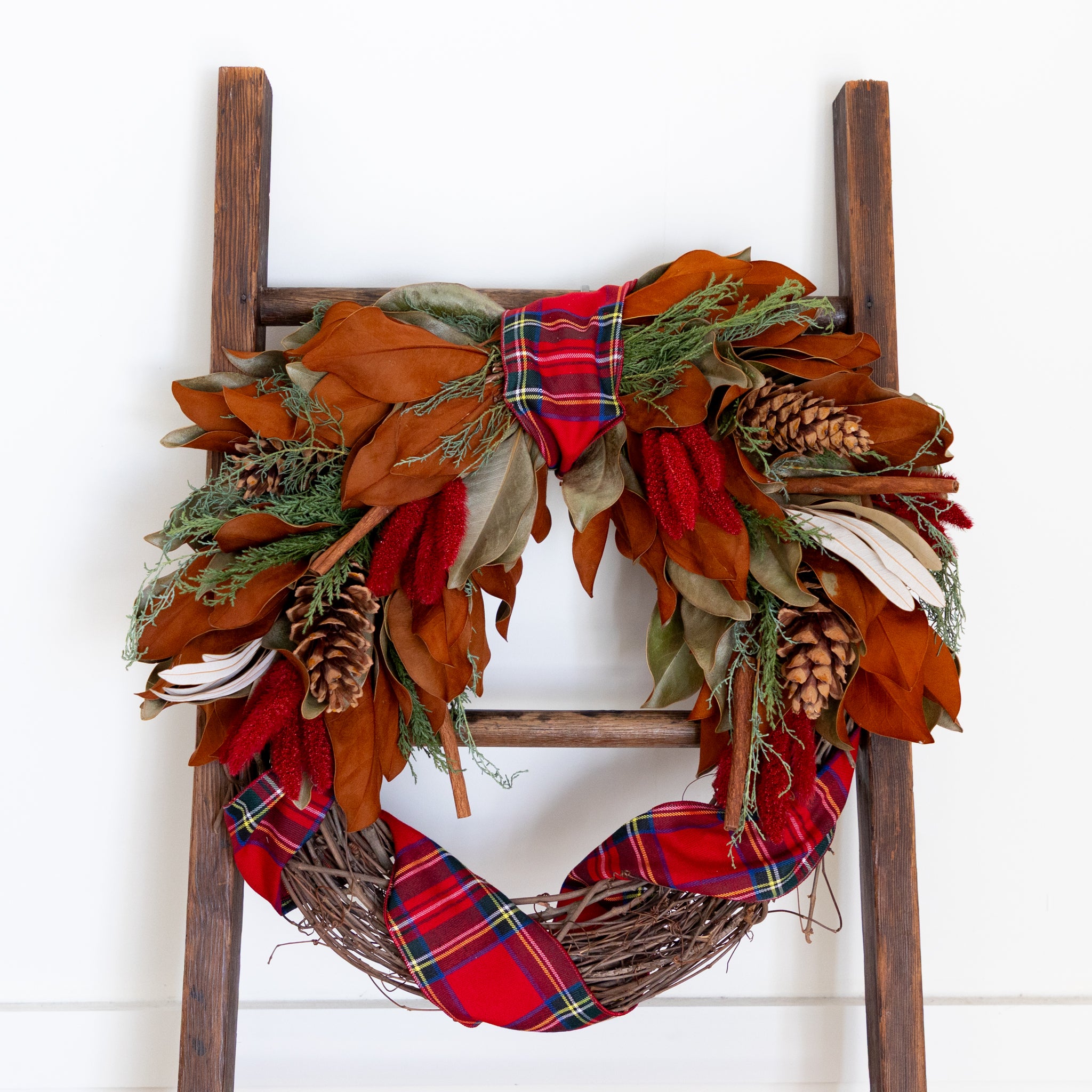 Decorative wreath with plaid ribbon and autumn leaves on a wooden ladder against a white background.
