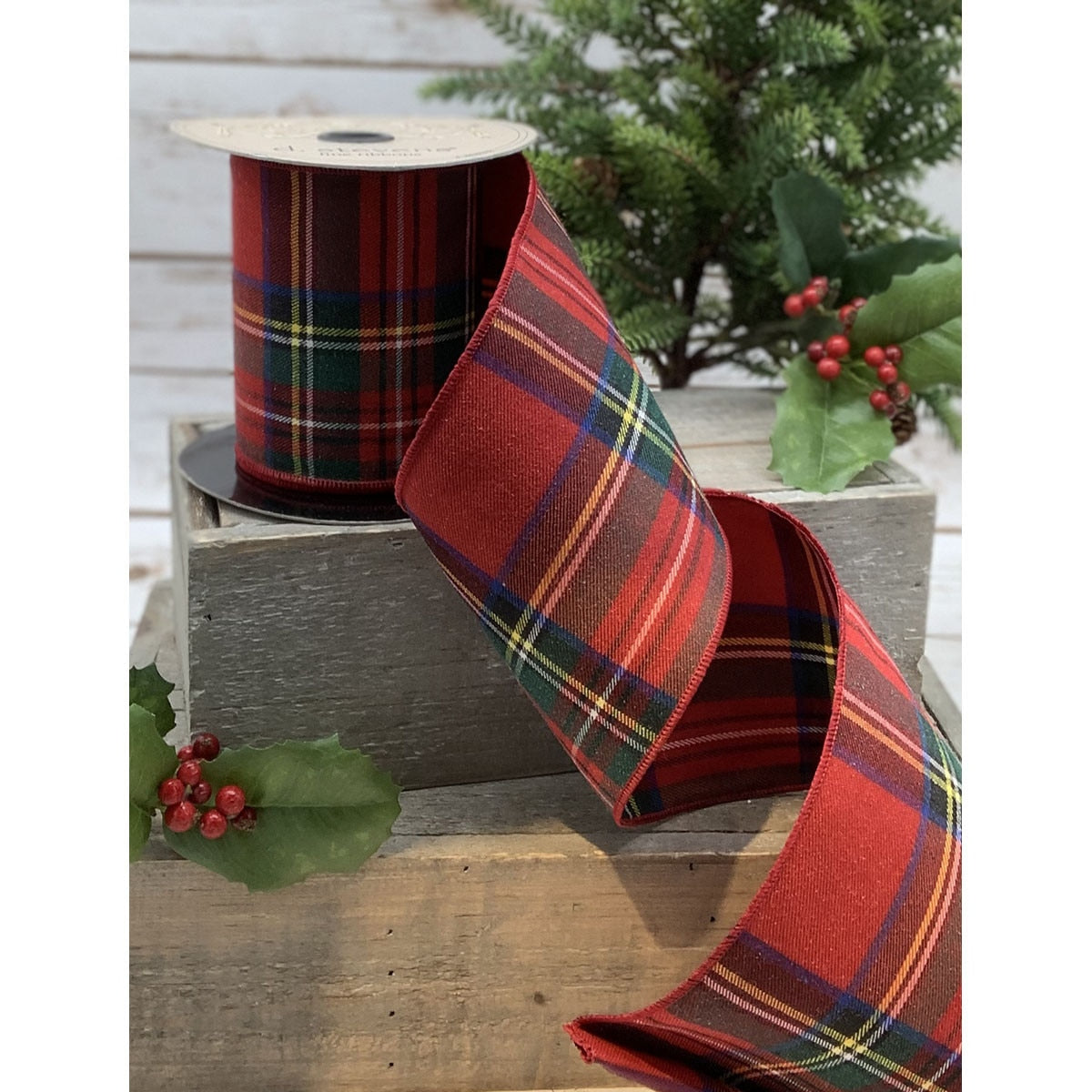 A roll of traditional red and green tartan ribbon placed on a wooden surface with decorative berries and green leaves in the background, indicating a festive setting.
