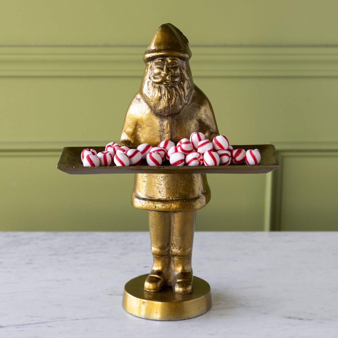 Gold statue of a Saint Nick holding a tray of peppermint candies against a green wall.