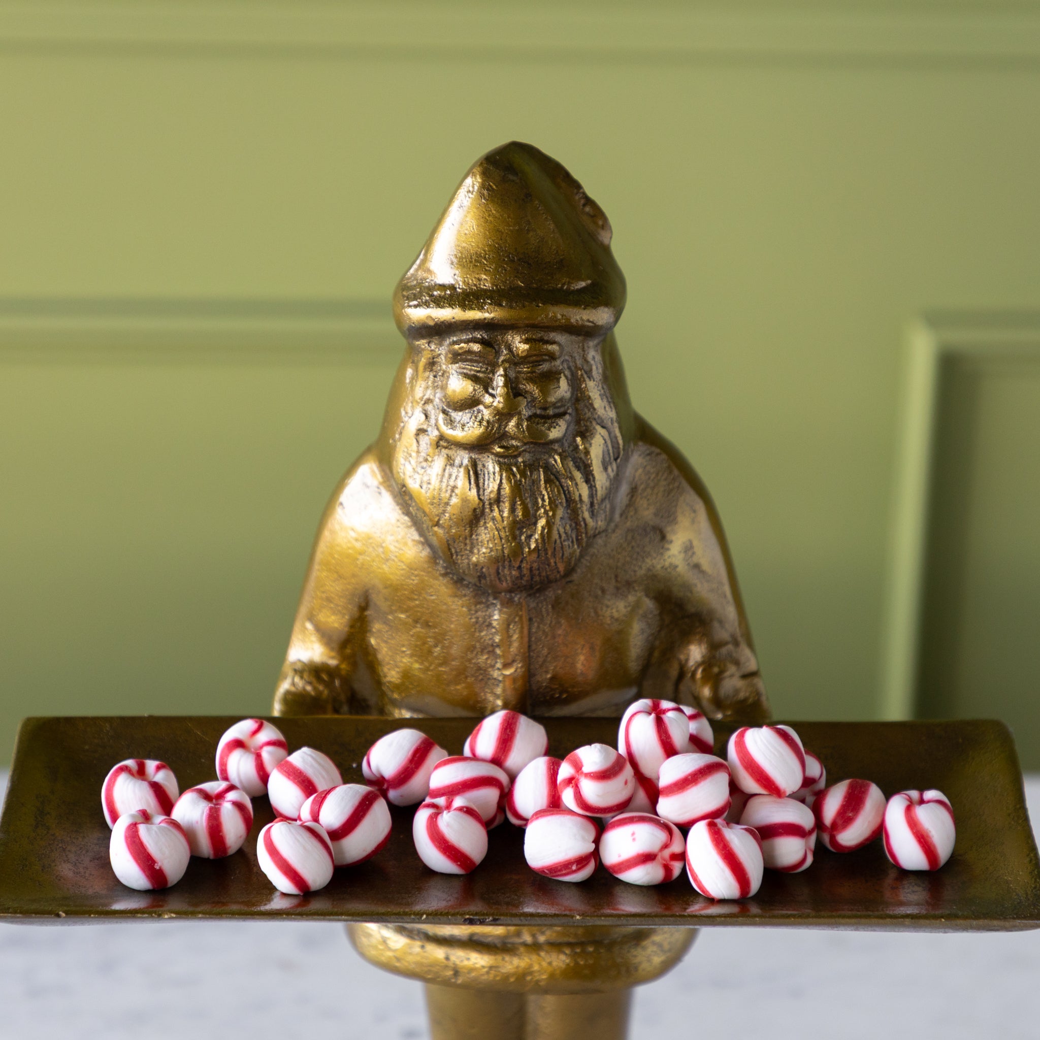 Gold statue of a Saint Nick holding a tray of peppermint candies against a green wall.