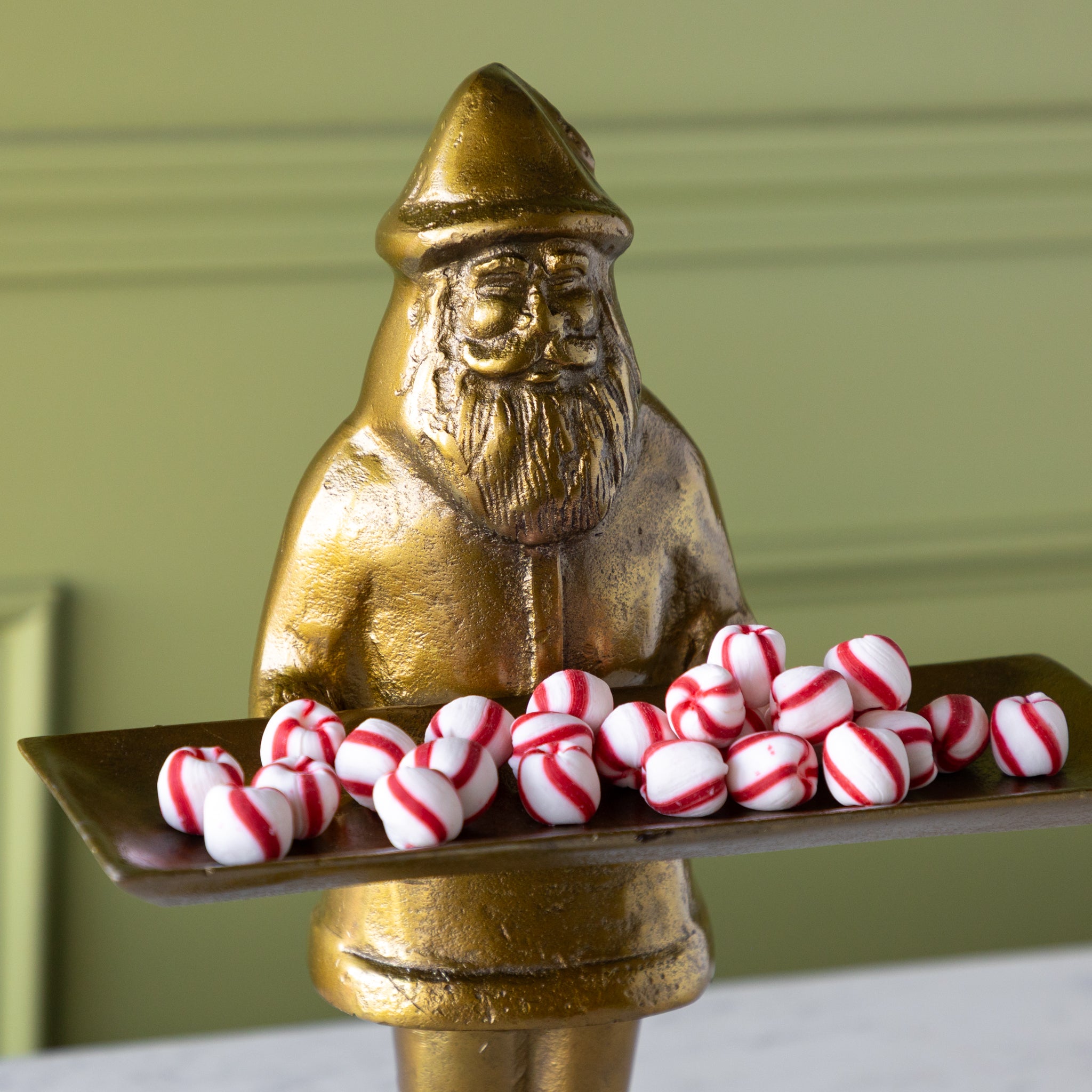 Gold statue of a Saint Nick holding a tray of peppermint candies against a green wall.