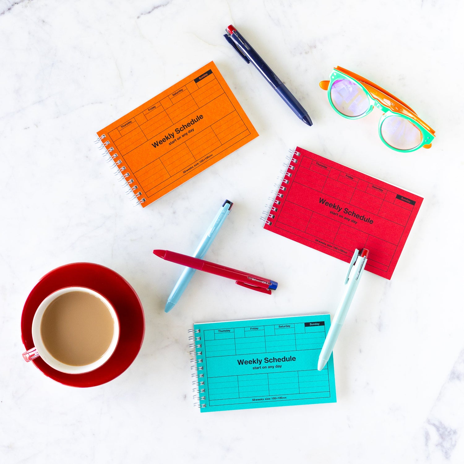 Colorful weekly schedule planners with pens, a cup of coffee, and glasses on a white surface.