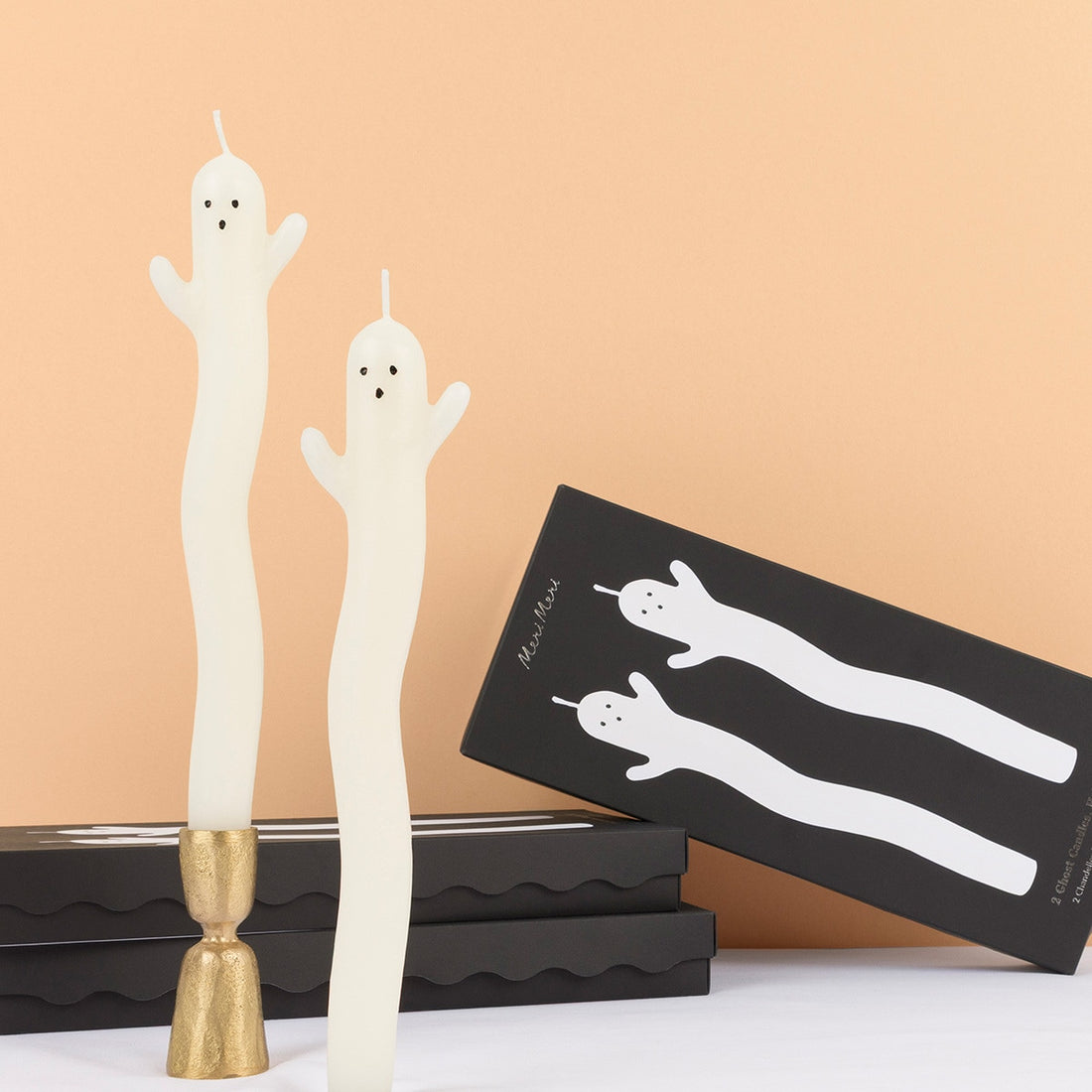 White ghost-shaped candles with gold candle holder and packaging on a beige background.