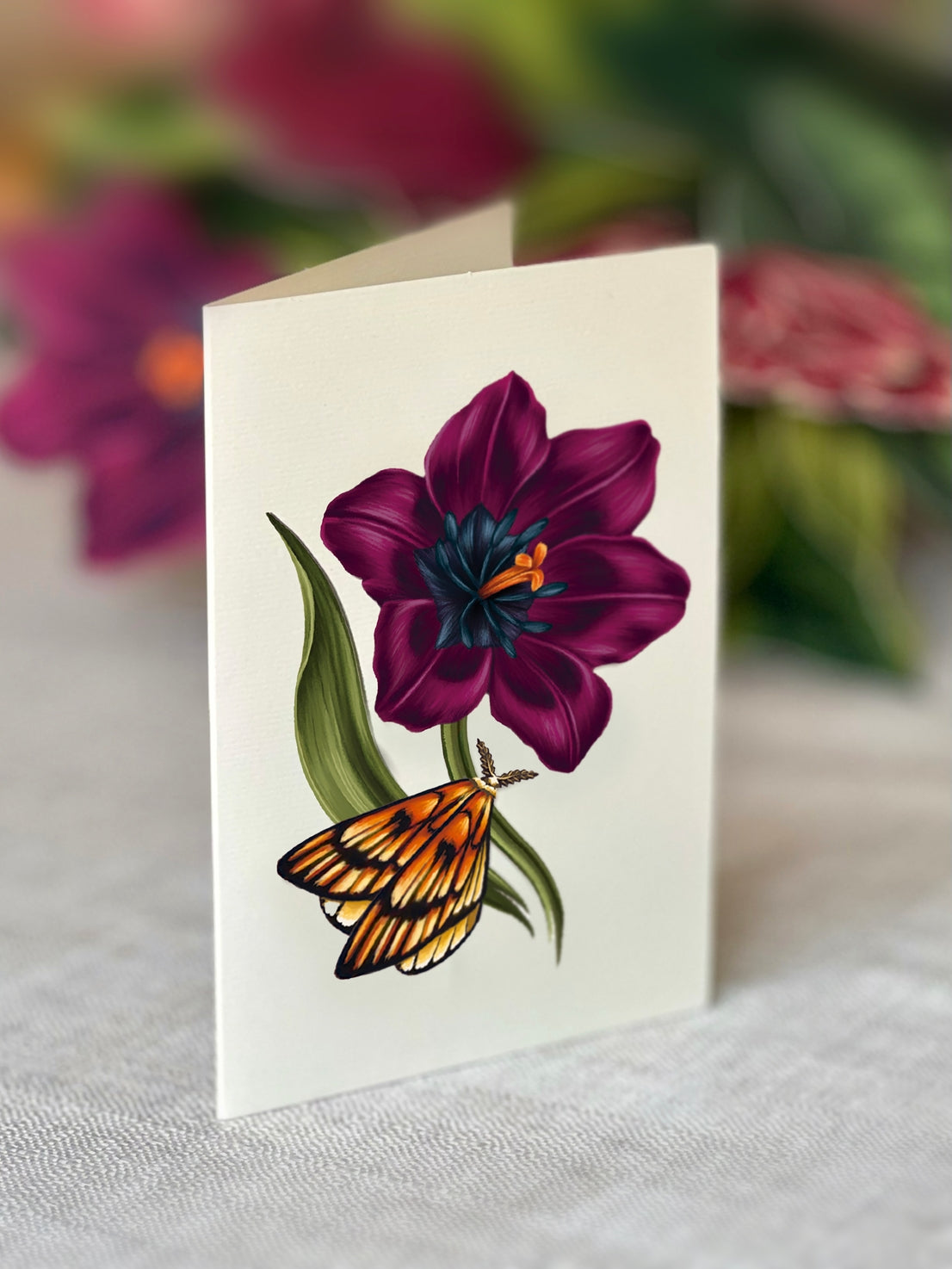 Greeting card with a purple flower and orange butterfly on a white surface with blurred flowers in the background