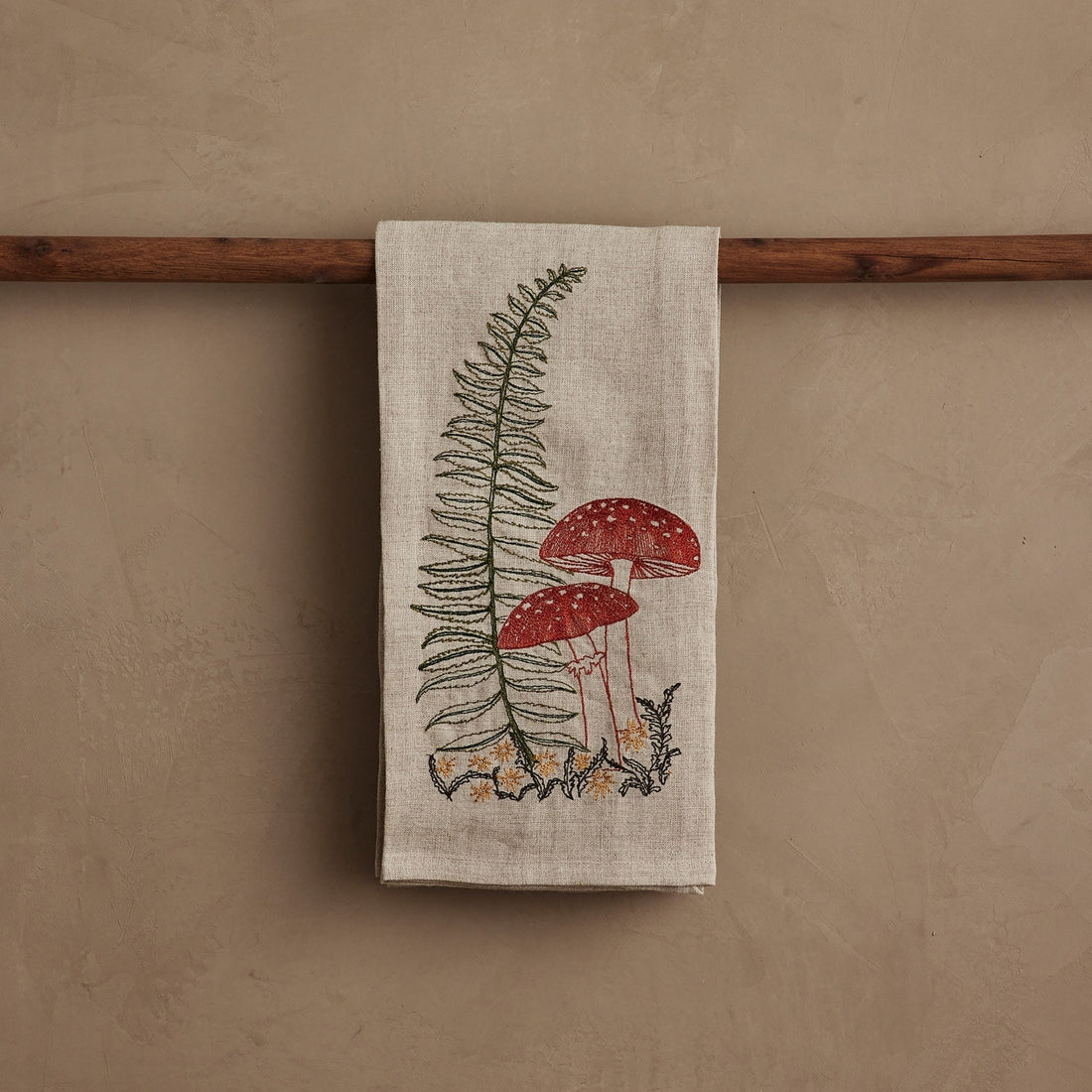 Tea towel with mushroom and fern design hanging on a wooden rod against a beige wall.