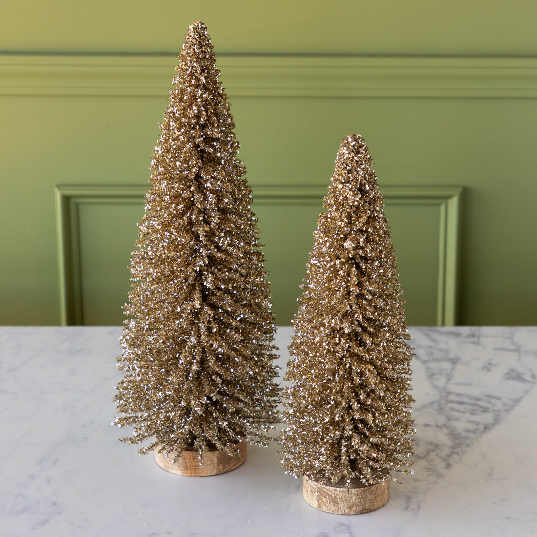 Two gold glittery bottlebrush trees on a marble surface with a green wall background.