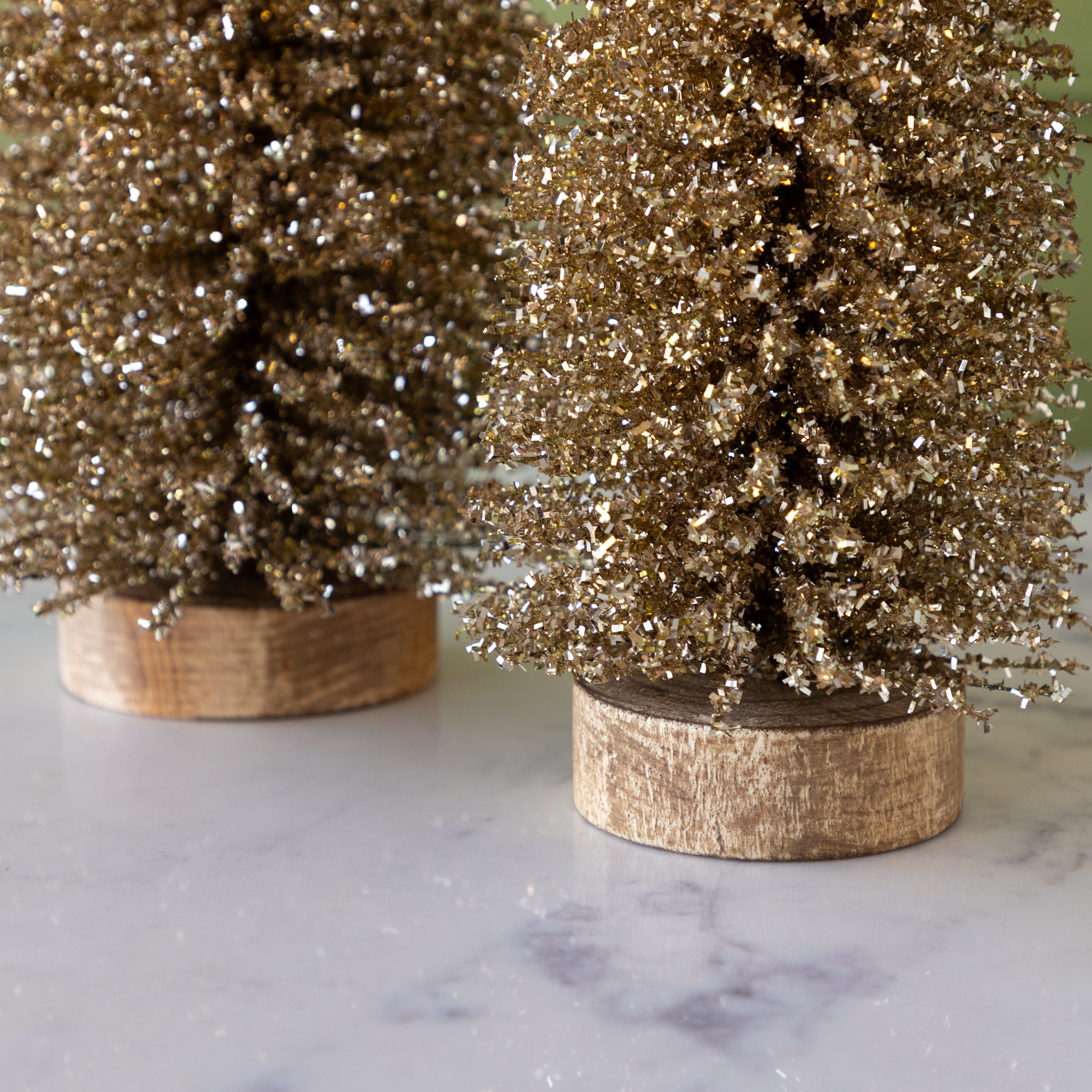 Two gold glittery bottlebrush trees on wooden bases, on a marble surface.