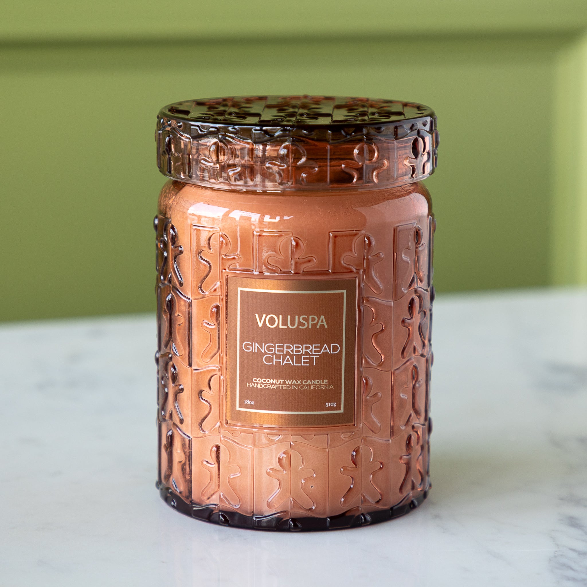 Decorative candle jar with intricate design on a marble surface against a green wall.