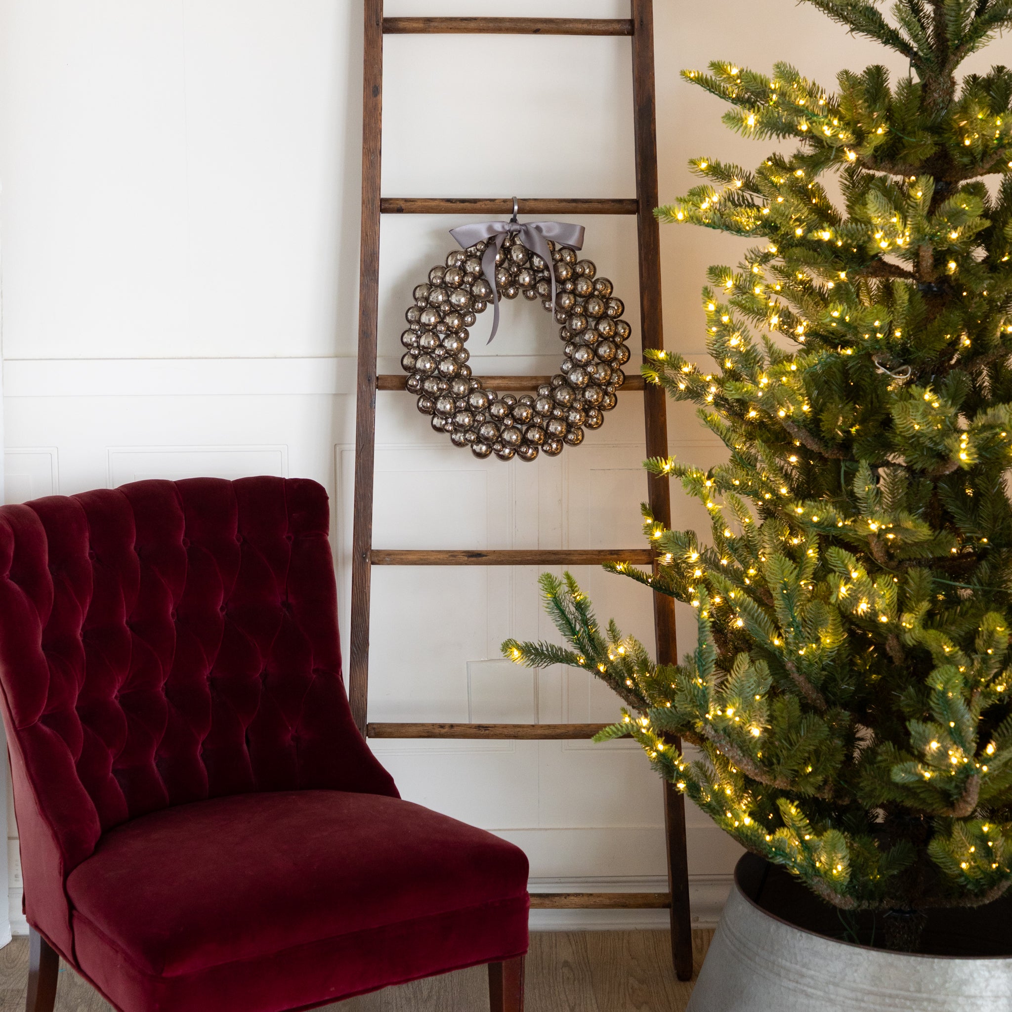 Antique Silver Bubble Wreath with a silver bow hanging on a wooden ladder next to a Christmas tree with lights and a red velvet chair, against a white wall.