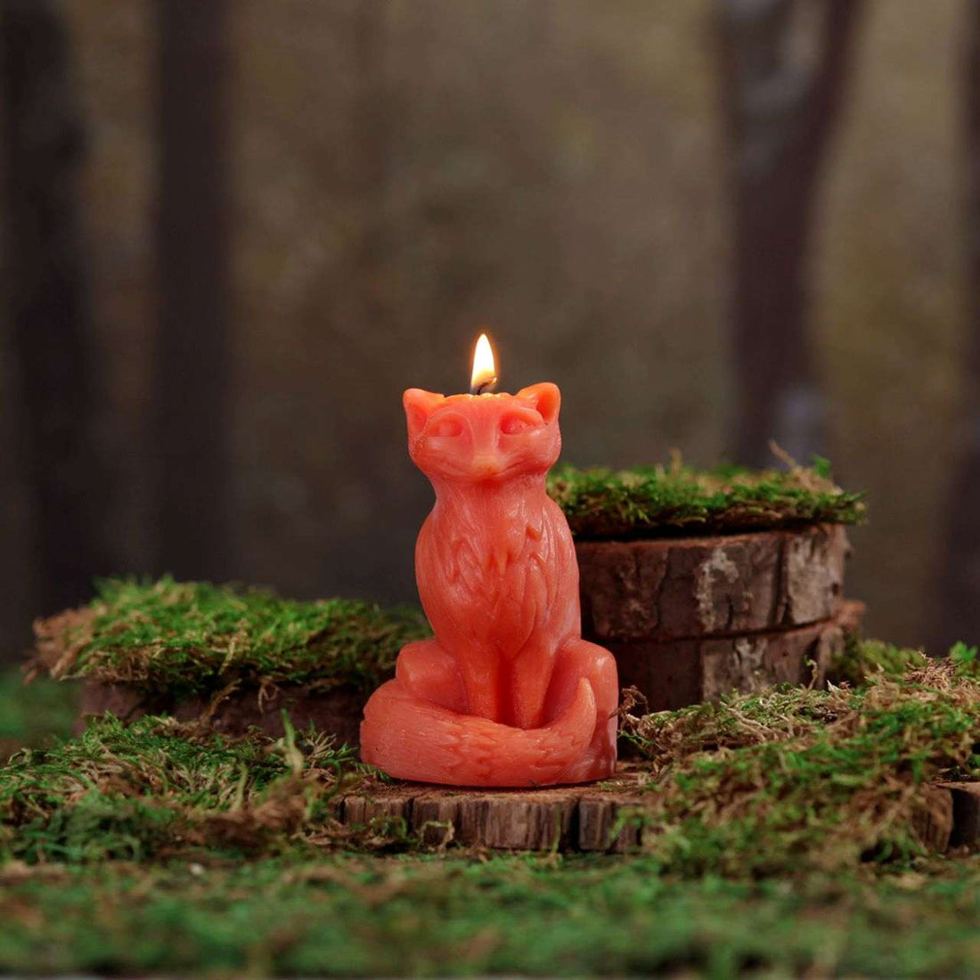 Fox-shaped candle with a lit flame on a wooden stump with grass, against a blurred natural background.