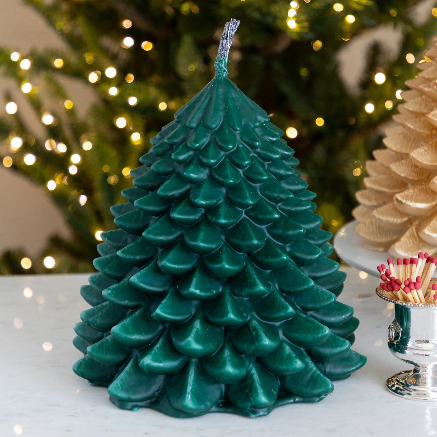 Green tree-shaped candle on a table with blurred Christmas lights in the background.