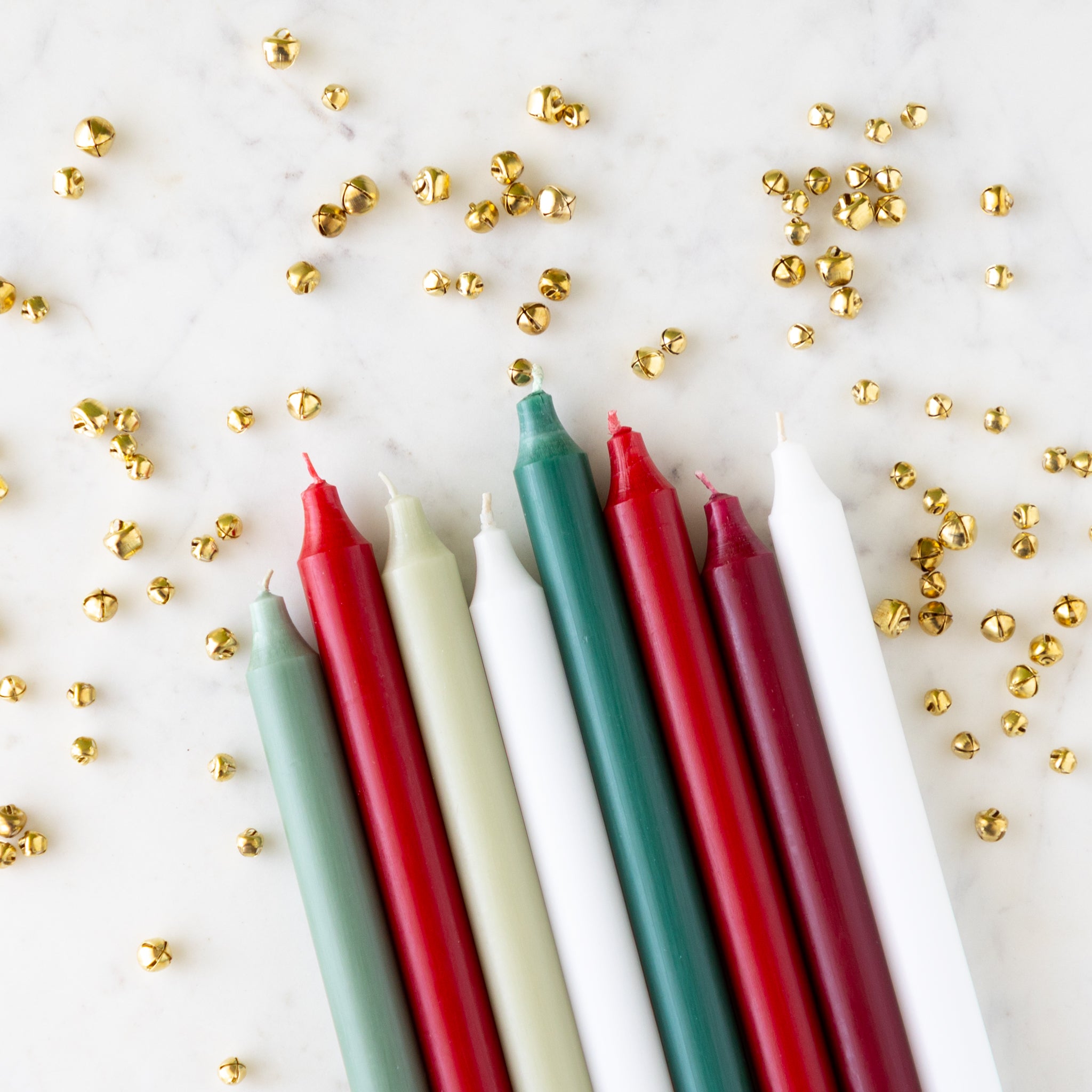 An assortment of holiday colored taper candles laying on a white background with small bells scattered.