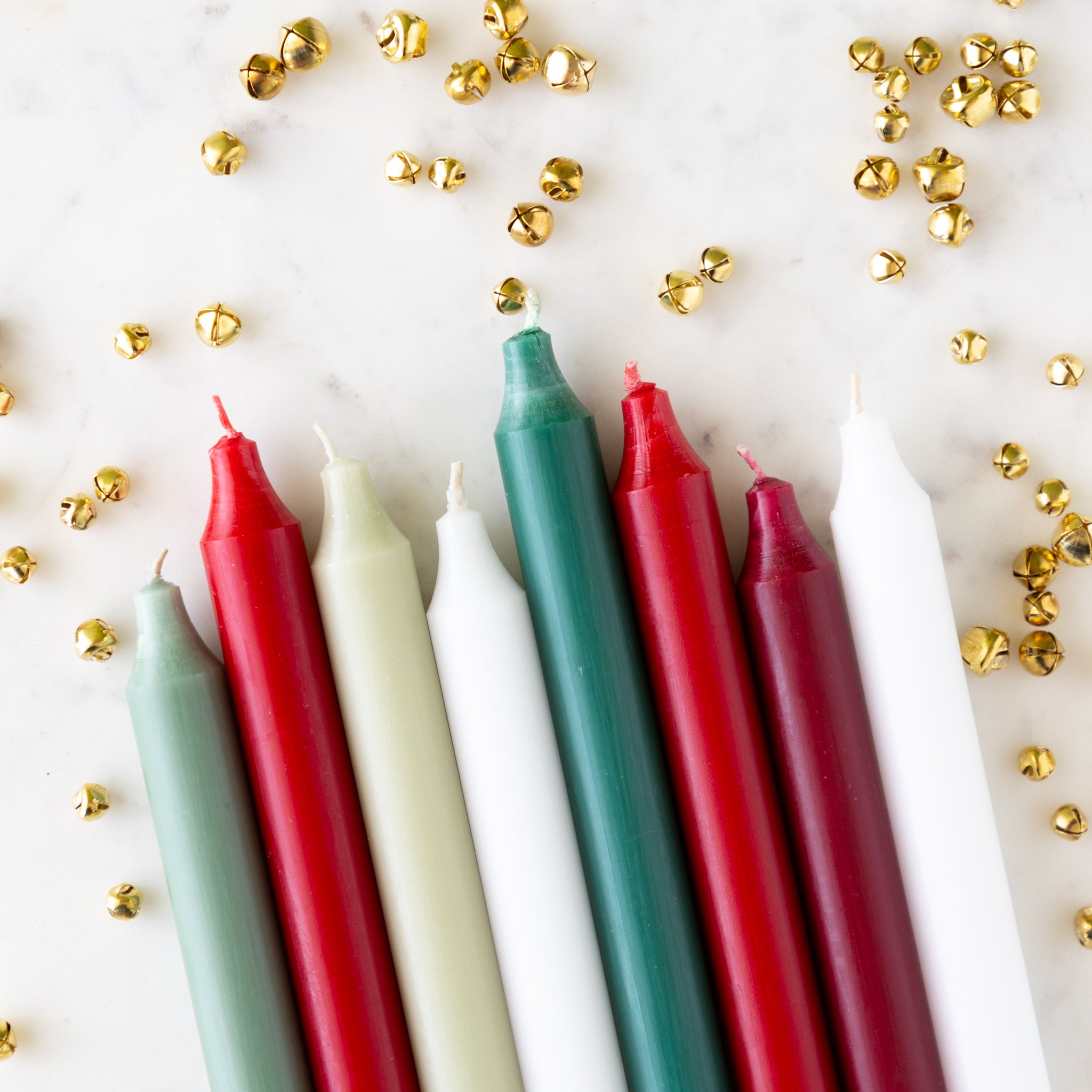 An assortment of holiday colored taper candles laying on a white background with small bells scattered.