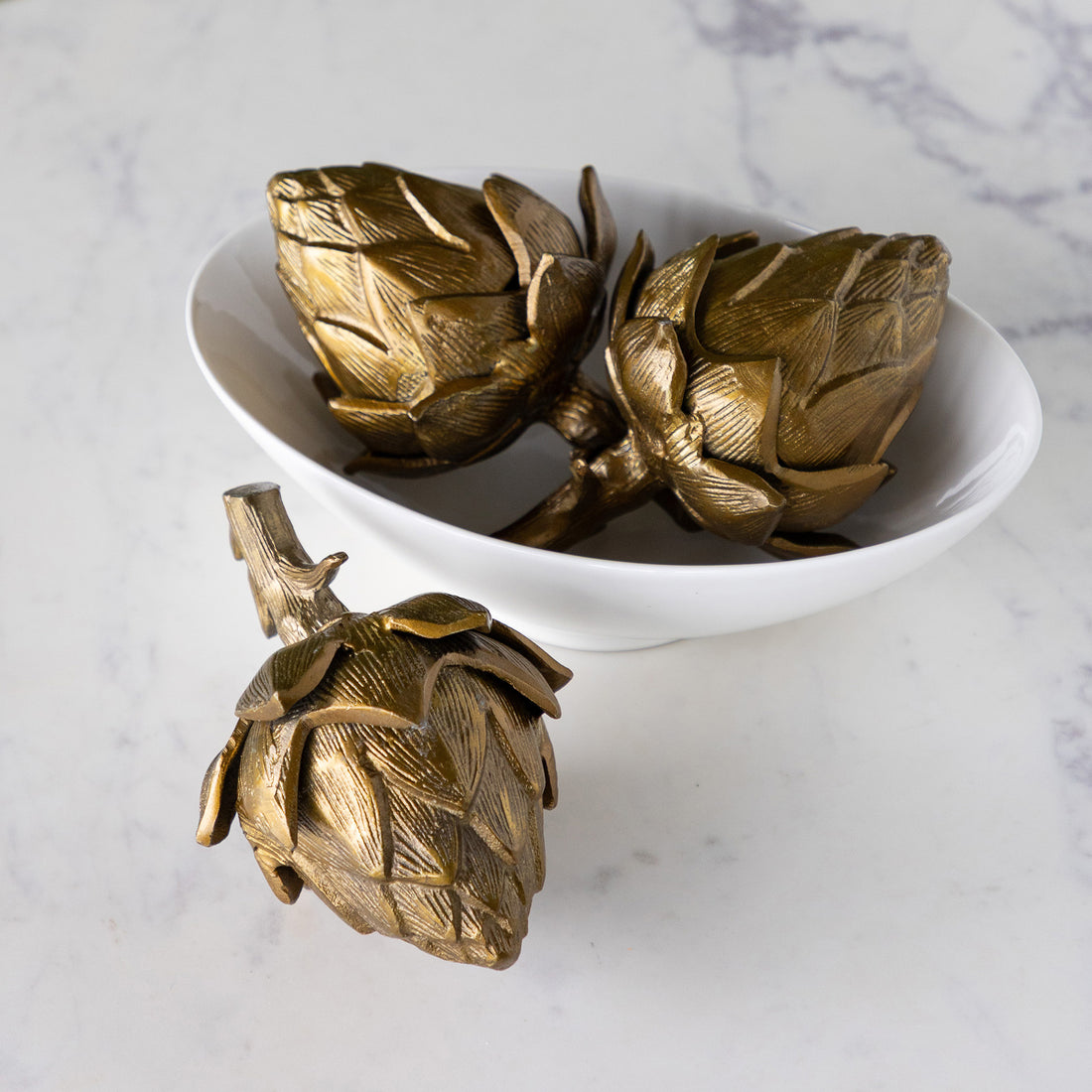 A bowl filled with two Gilded Artichokes and one outside of the bowl on a marble table.
