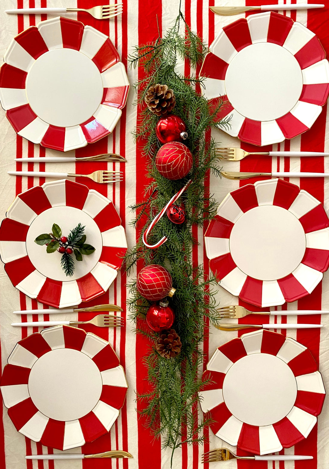 A set of red and white scalloped paper plates with gold foil edges, arranged on a table to mimic a festive dinner setting, decorated with candy canes, pine branches, and holiday ornaments.