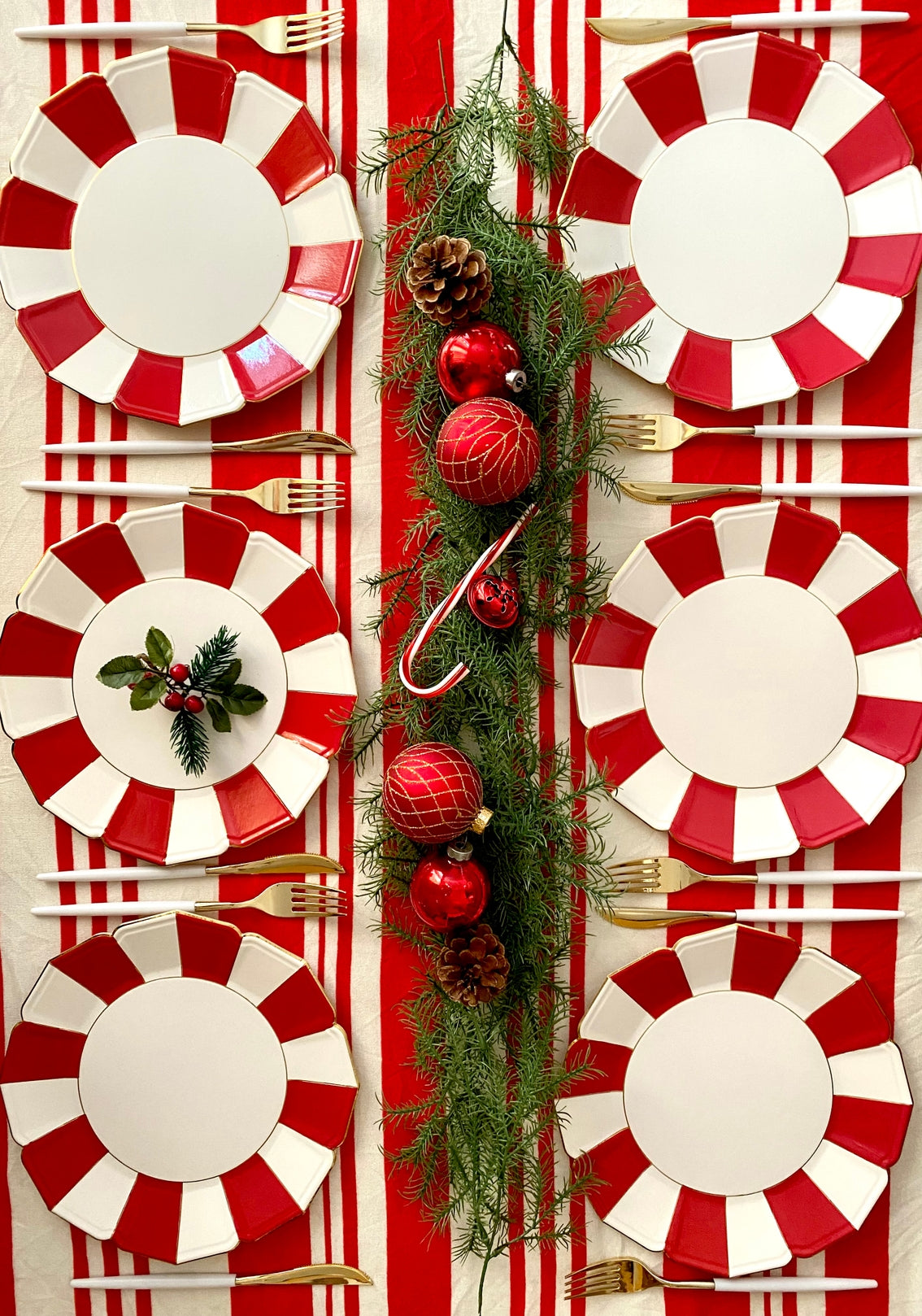 A set of red and white scalloped paper plates with gold foil edges, arranged on a table to mimic a festive dinner setting, decorated with candy canes, pine branches, and holiday ornaments.