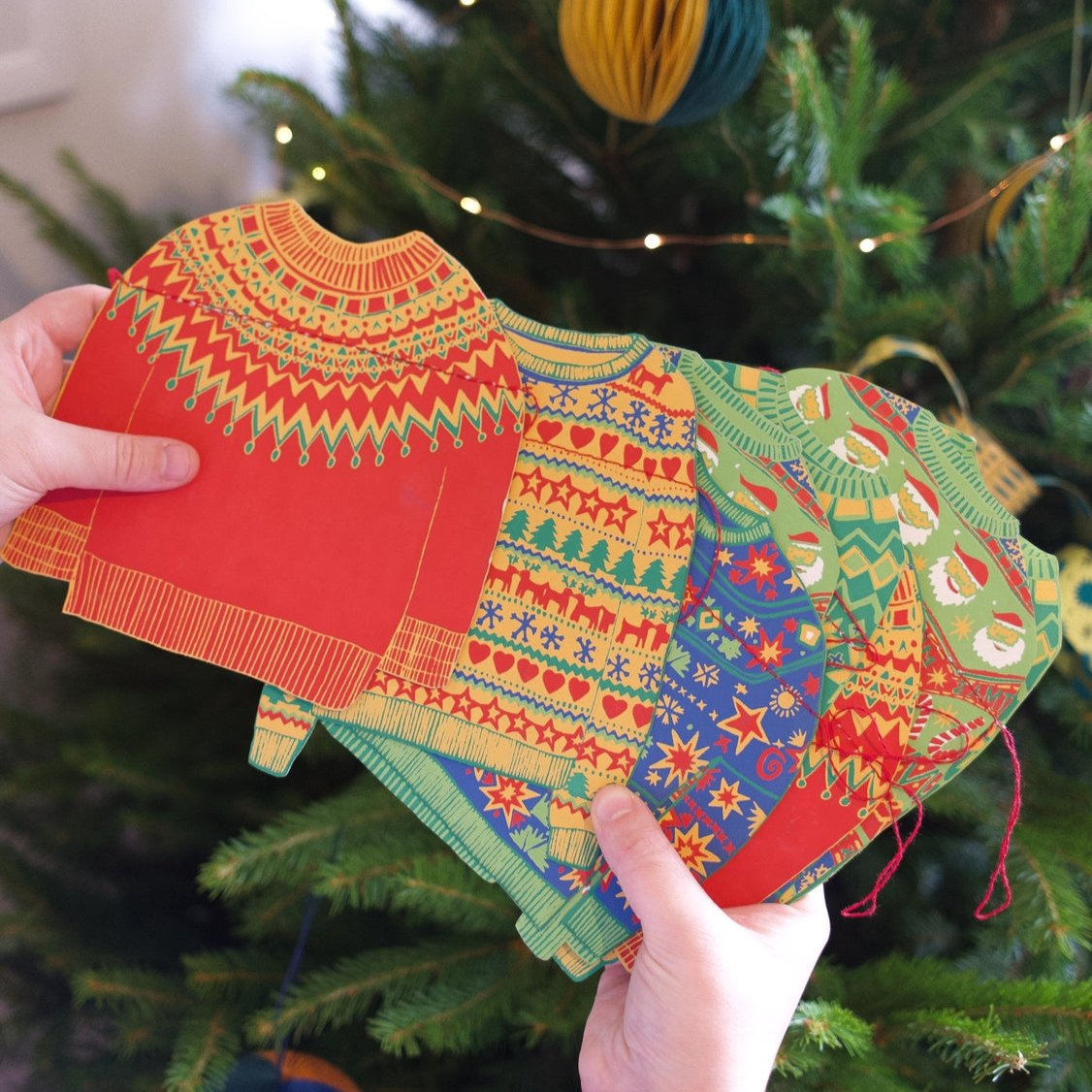 A pair of hands holding the paper sweaters fanned out to show the various festive designs.