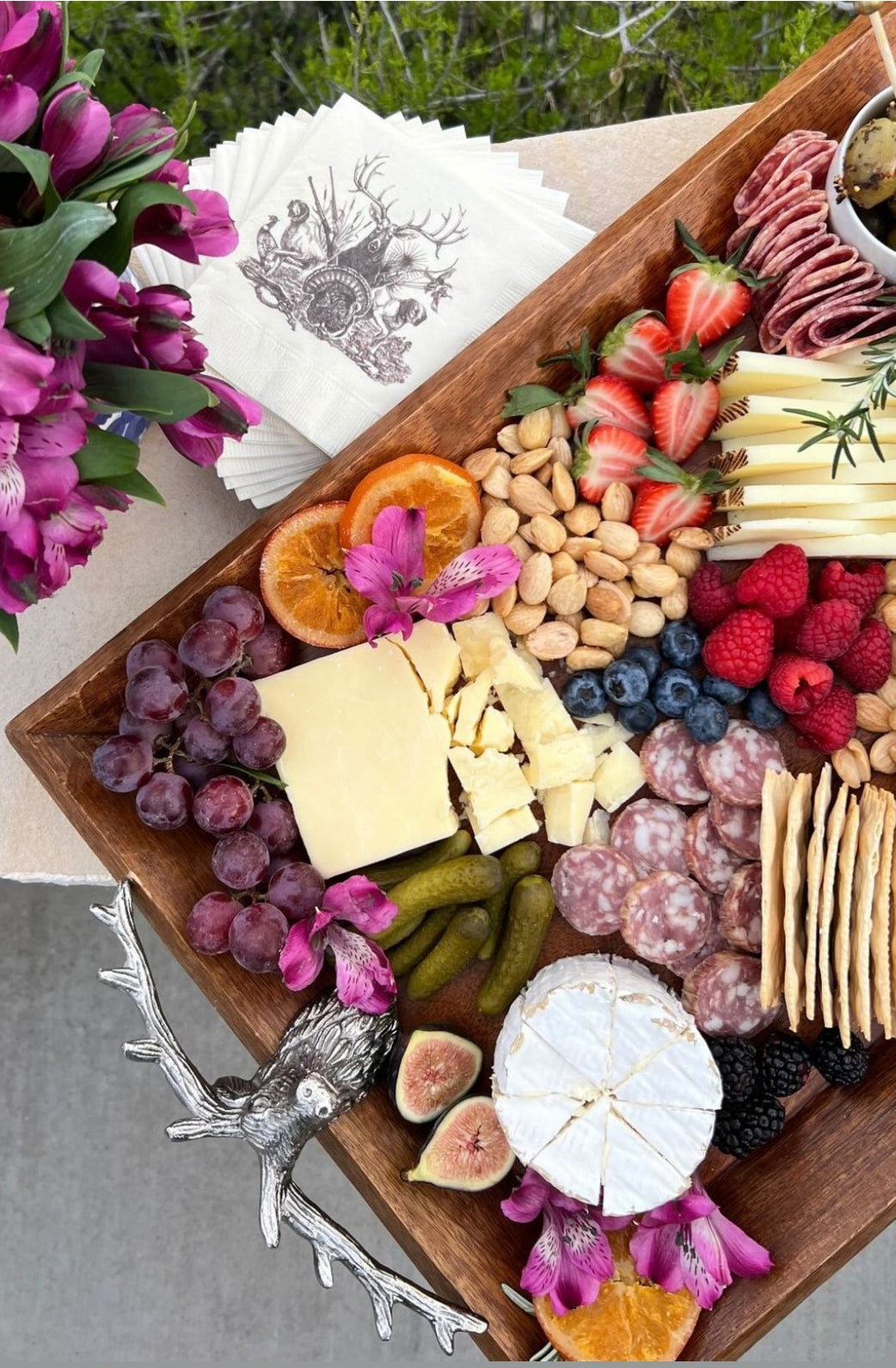 Wooden charcuterie board with assorted meats, cheeses, fruits, and crackers on a table with flowers and On the Hunt napkins.