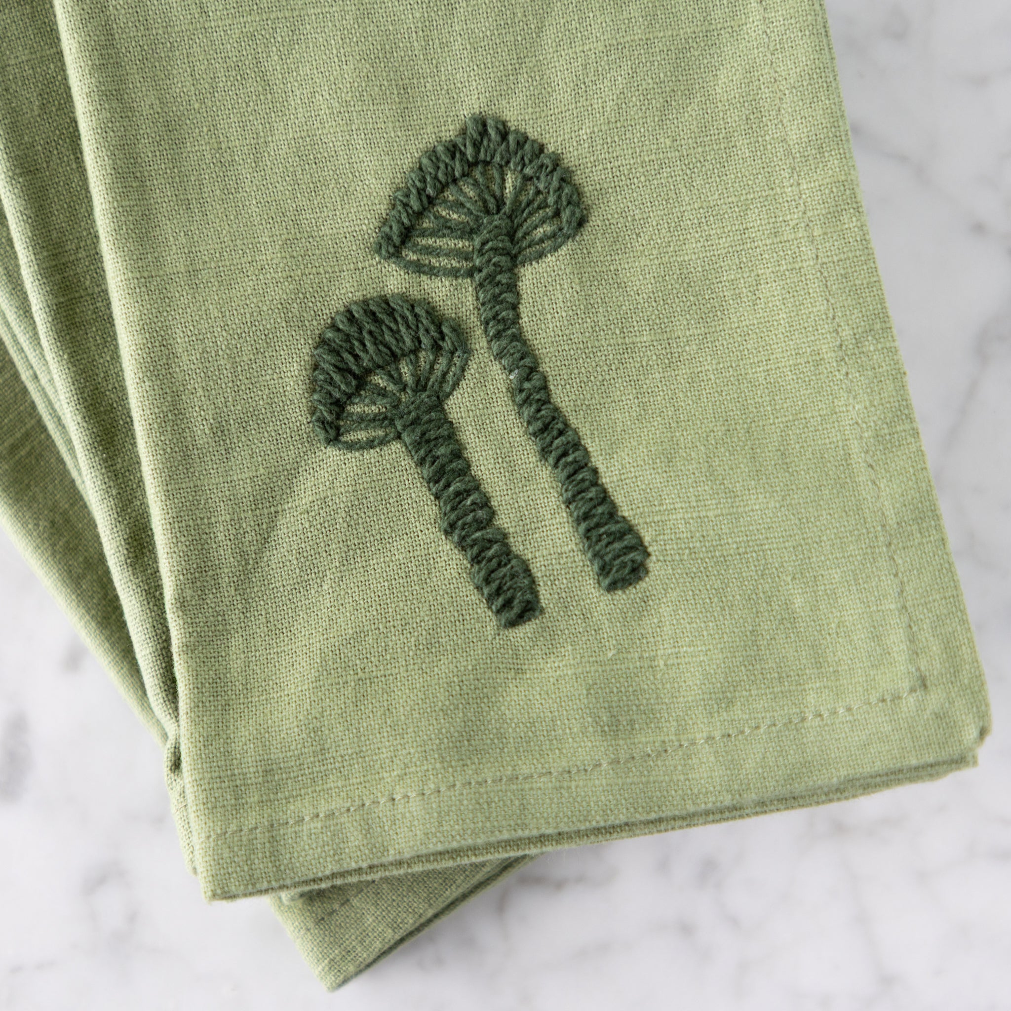 Green embroidered napkin with mushroom design on a marble surface.