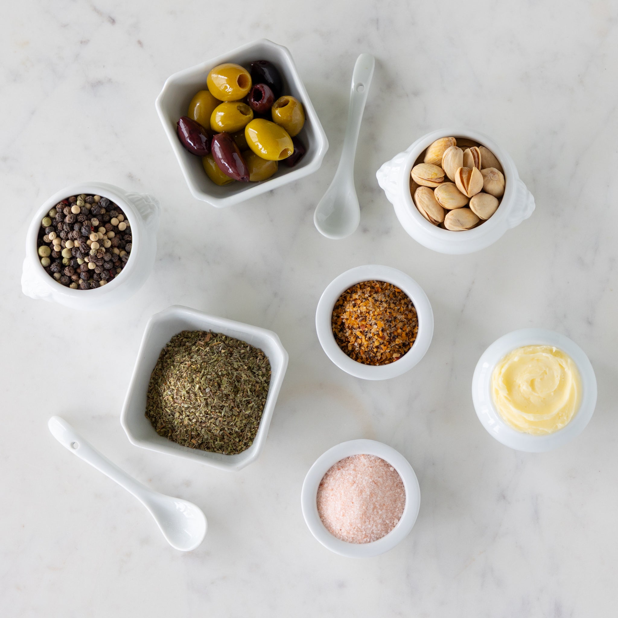 Assorted spices and seasonings in small white bowls next to white spoons on a marble surface.
