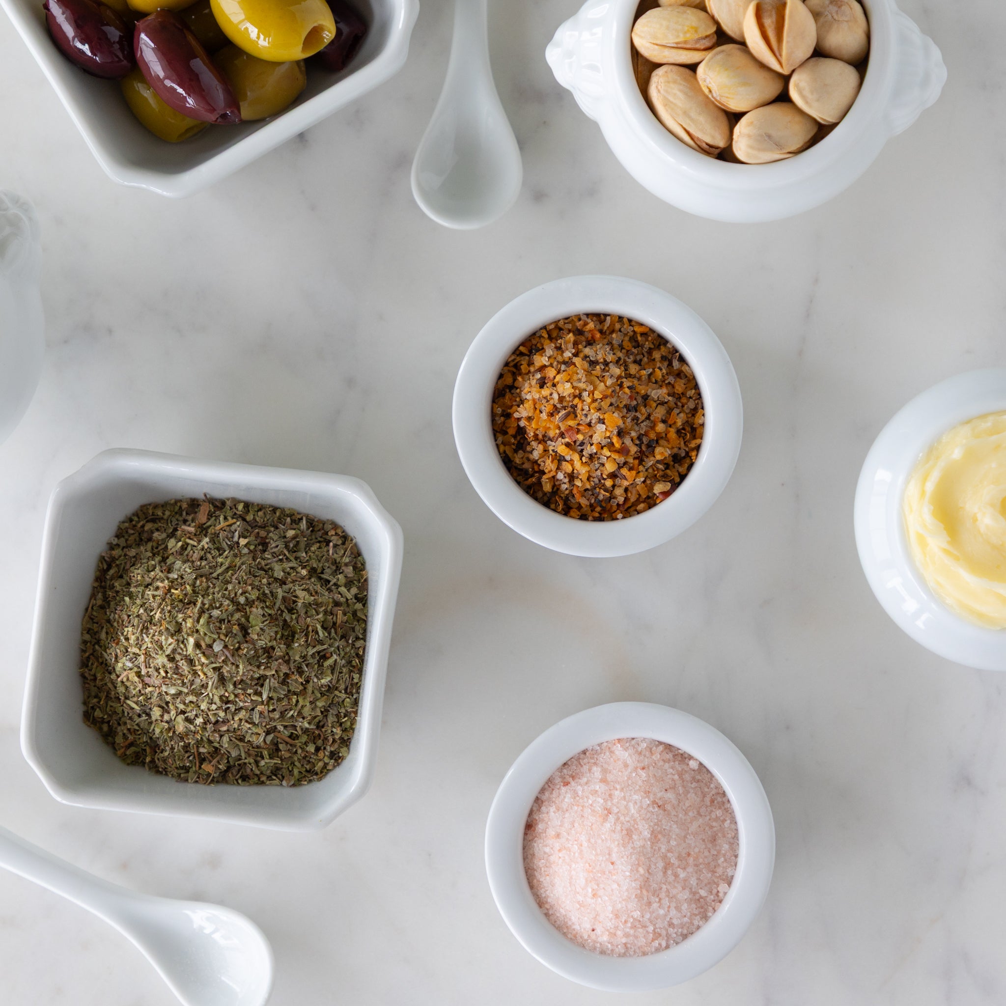 Assorted spices and seasonings in small white bowls next to white spoons on a marble surface.