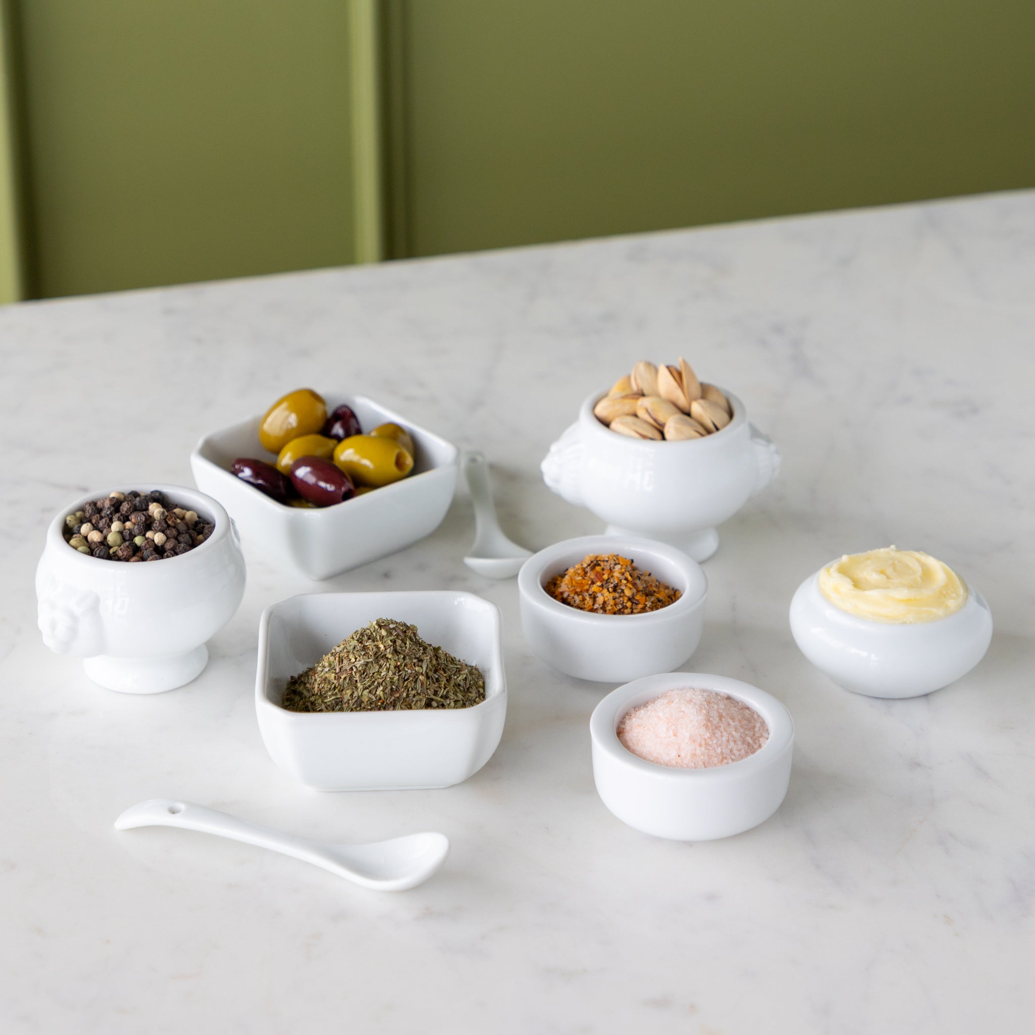 Assorted spices and seasonings in small white bowls next to white spoons on a marble surface.