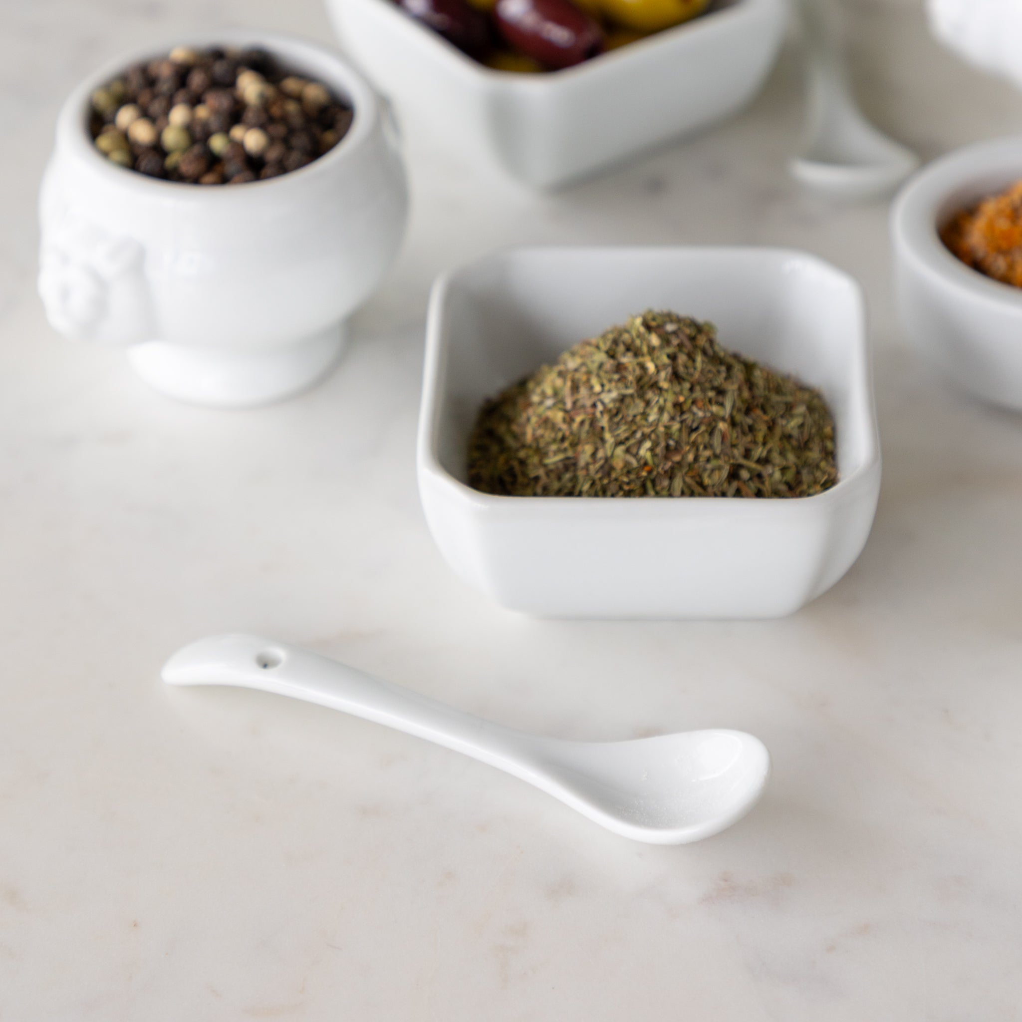 White bowls with various spices and a white spoon on a light surface.