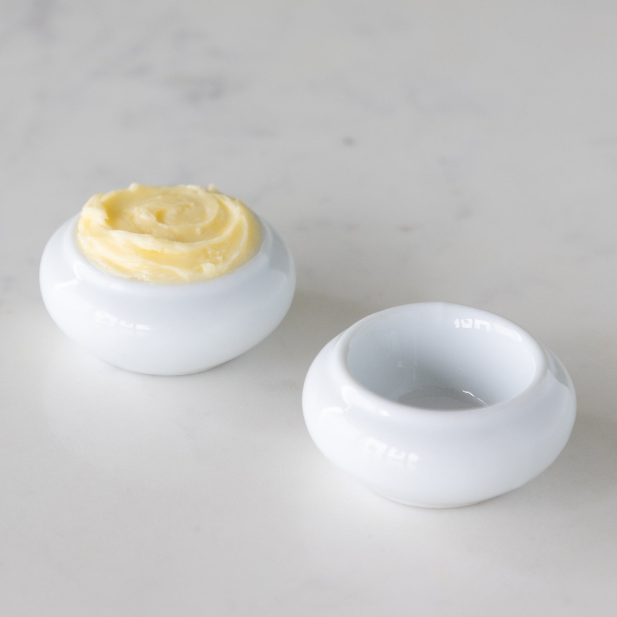 Two white ceramic butter bots with one containing butter and another empty, on a marble table.