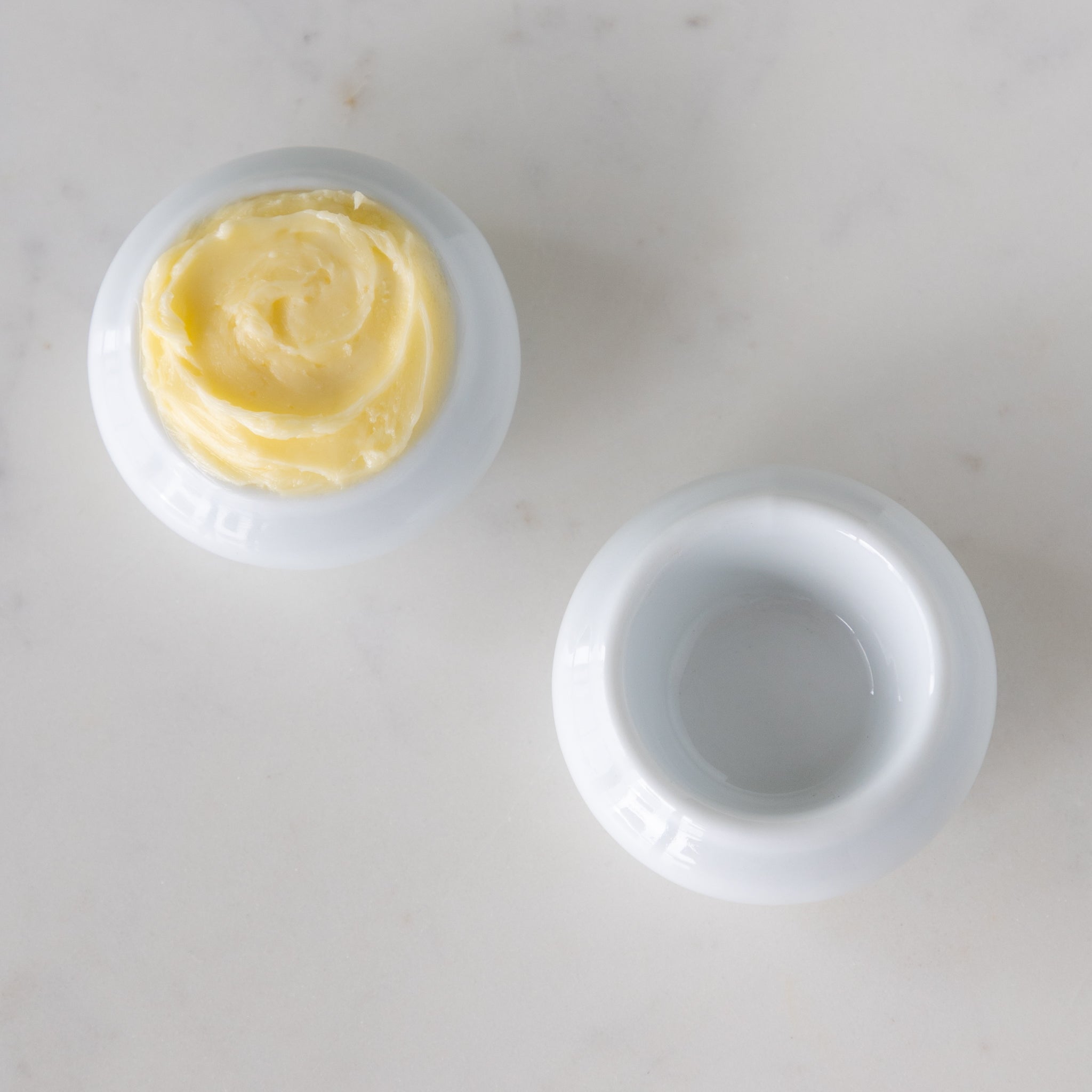 Two white ceramic butter bots with one containing butter and another empty, on a marble table.