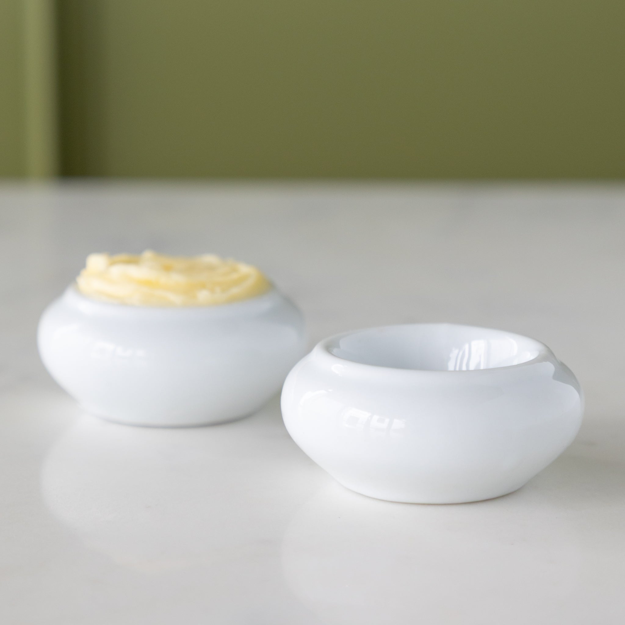 Two white ceramic butter bots with one containing butter and another empty, on a marble table.