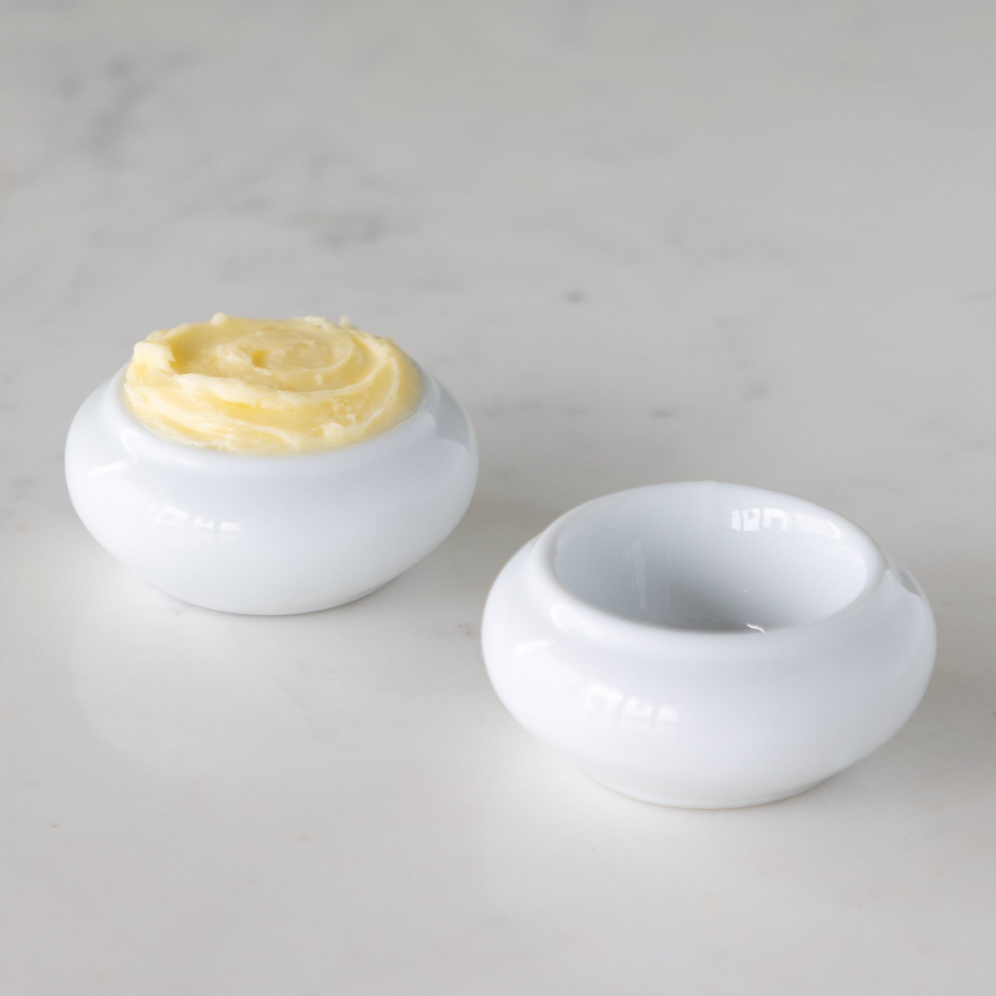Two white ceramic butter bots with one containing butter and another empty, on a marble table.