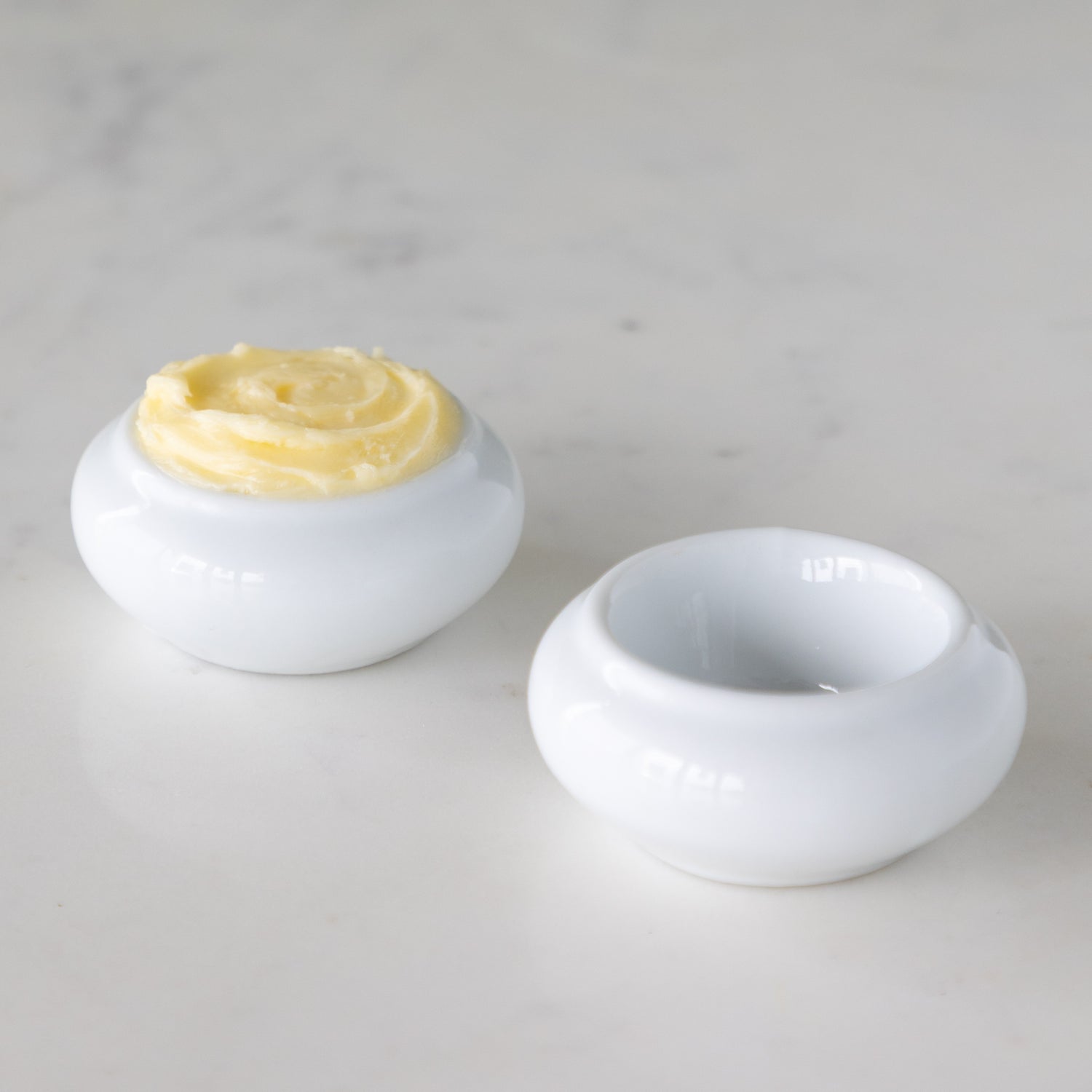 Two white ceramic butter bots with one containing butter and another empty, on a marble table. 