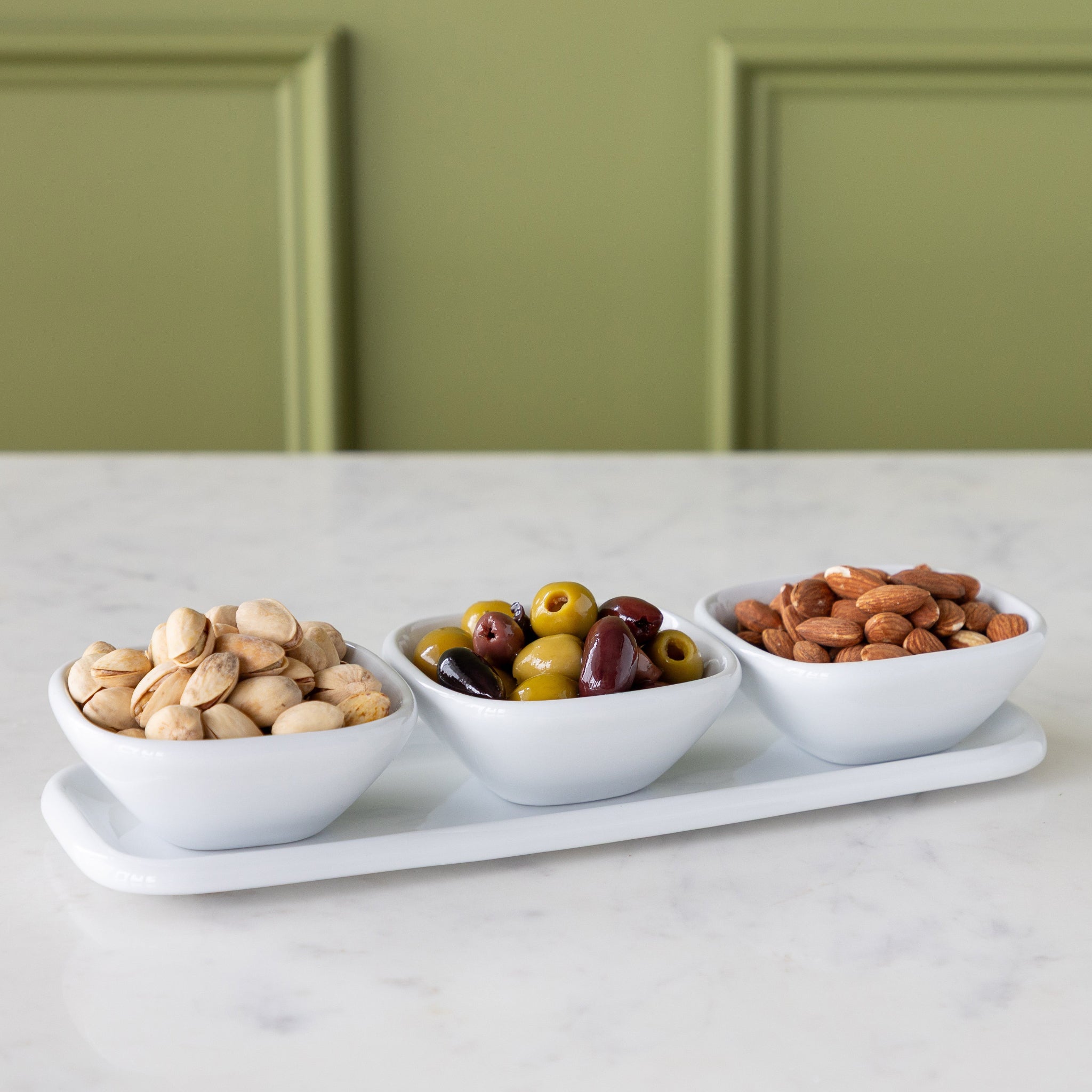 Three white bowls with nuts and olives on a tray with a green wall background.