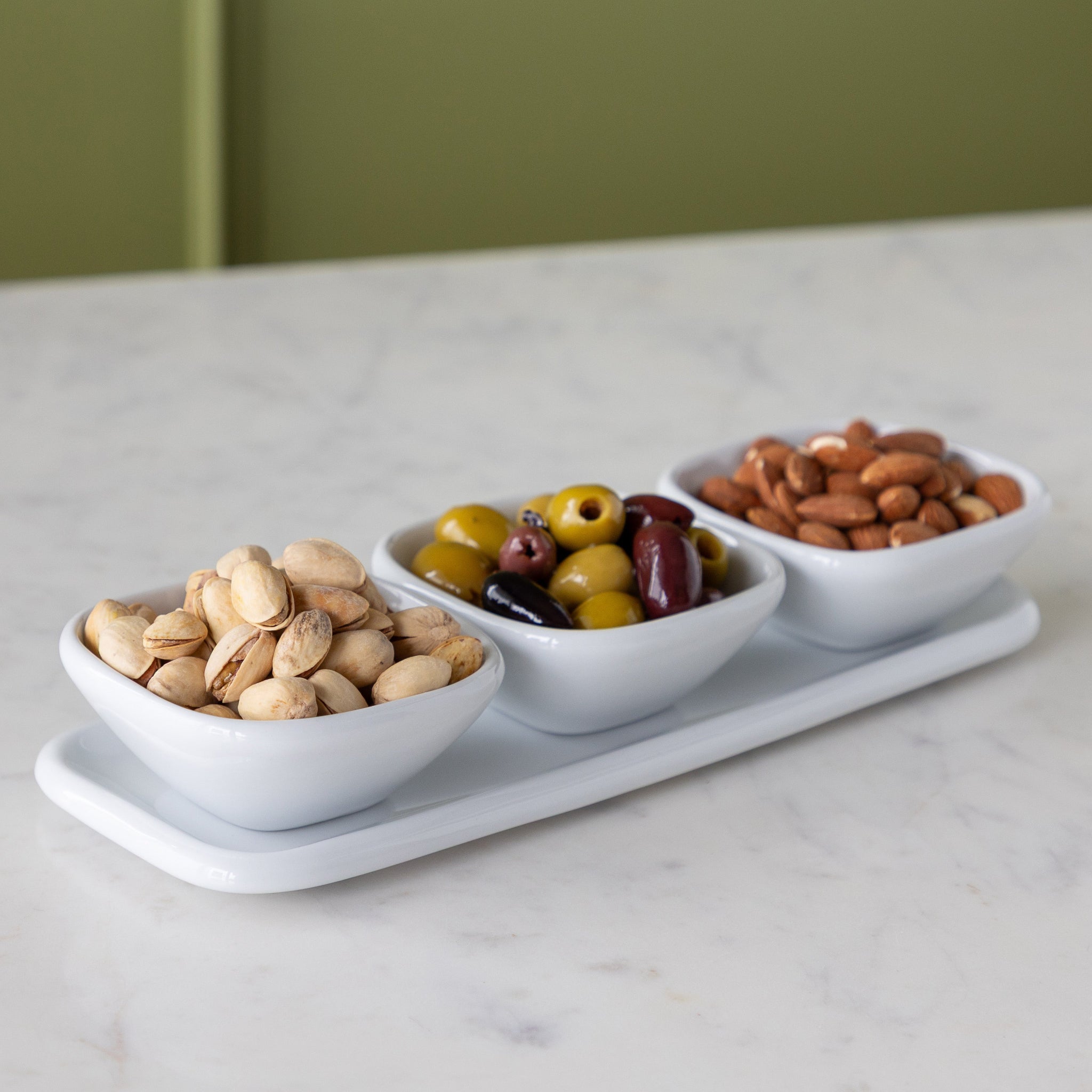 Three white bowls on a tray with nuts and olives on a marble surface.