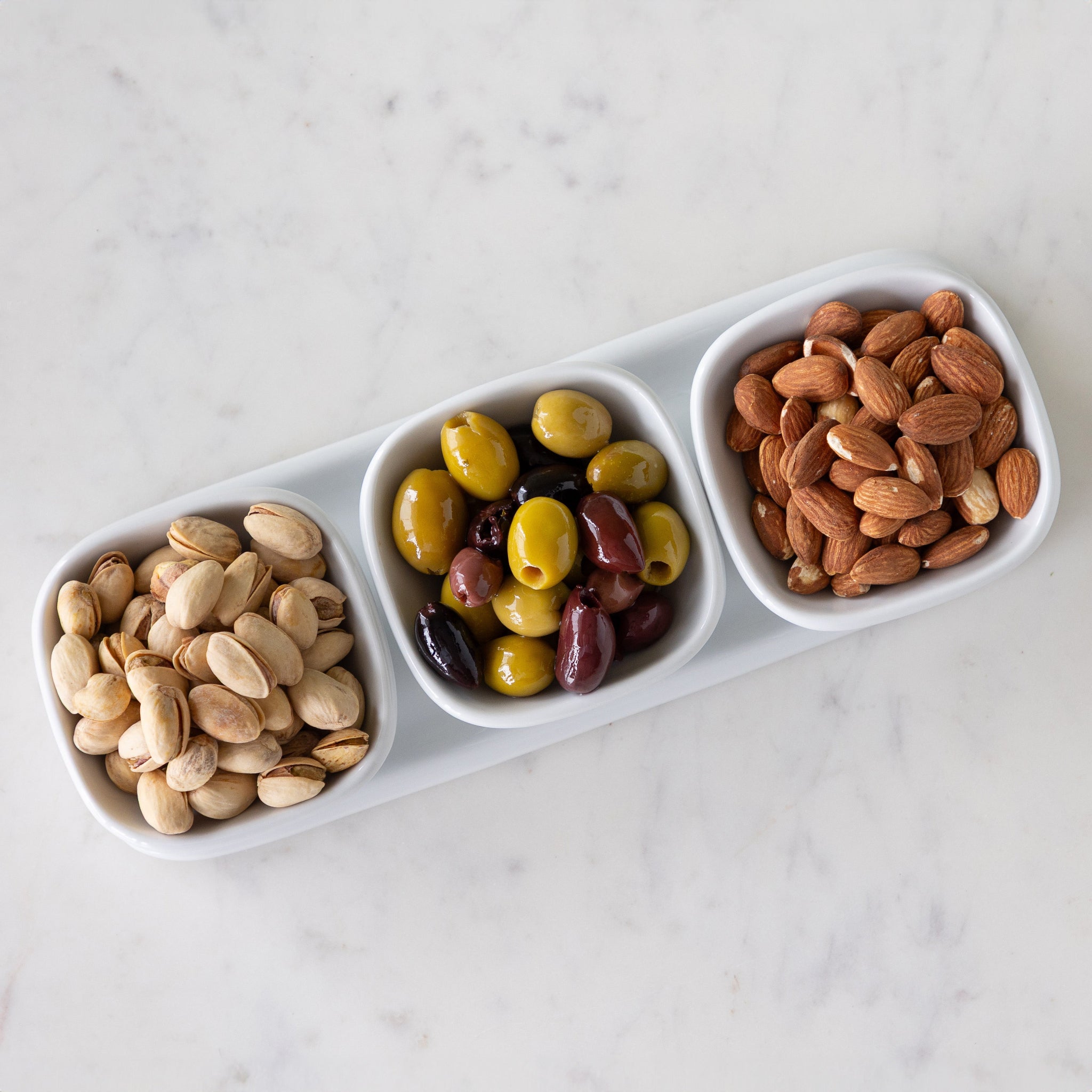 Three white porcelain bowls filled with pistachios, olives and almonds on a porcelain tray.