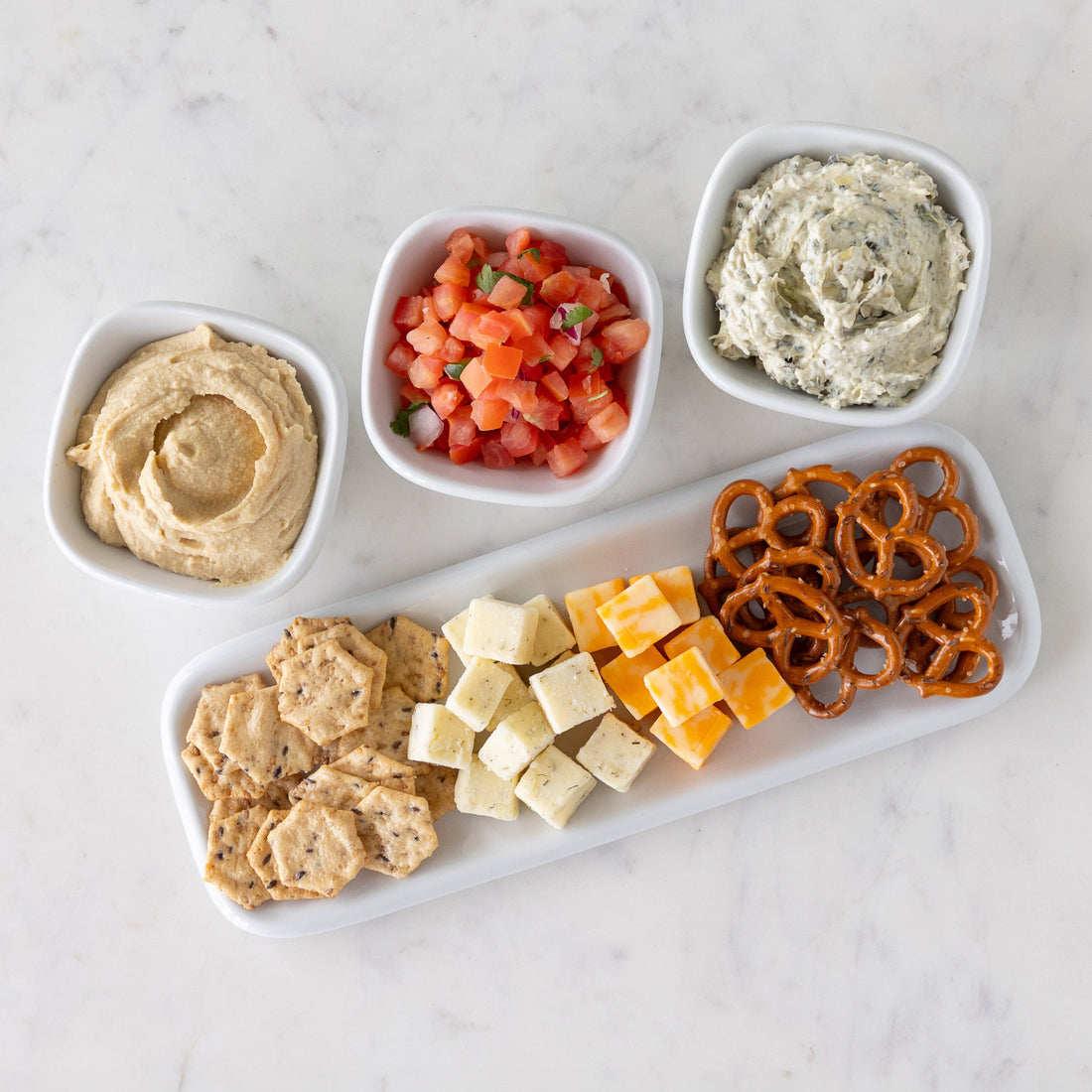 Assorted dips in white bowls and assorted snacks including crackers, cheese cubes, and pretzels on a white marble tray.