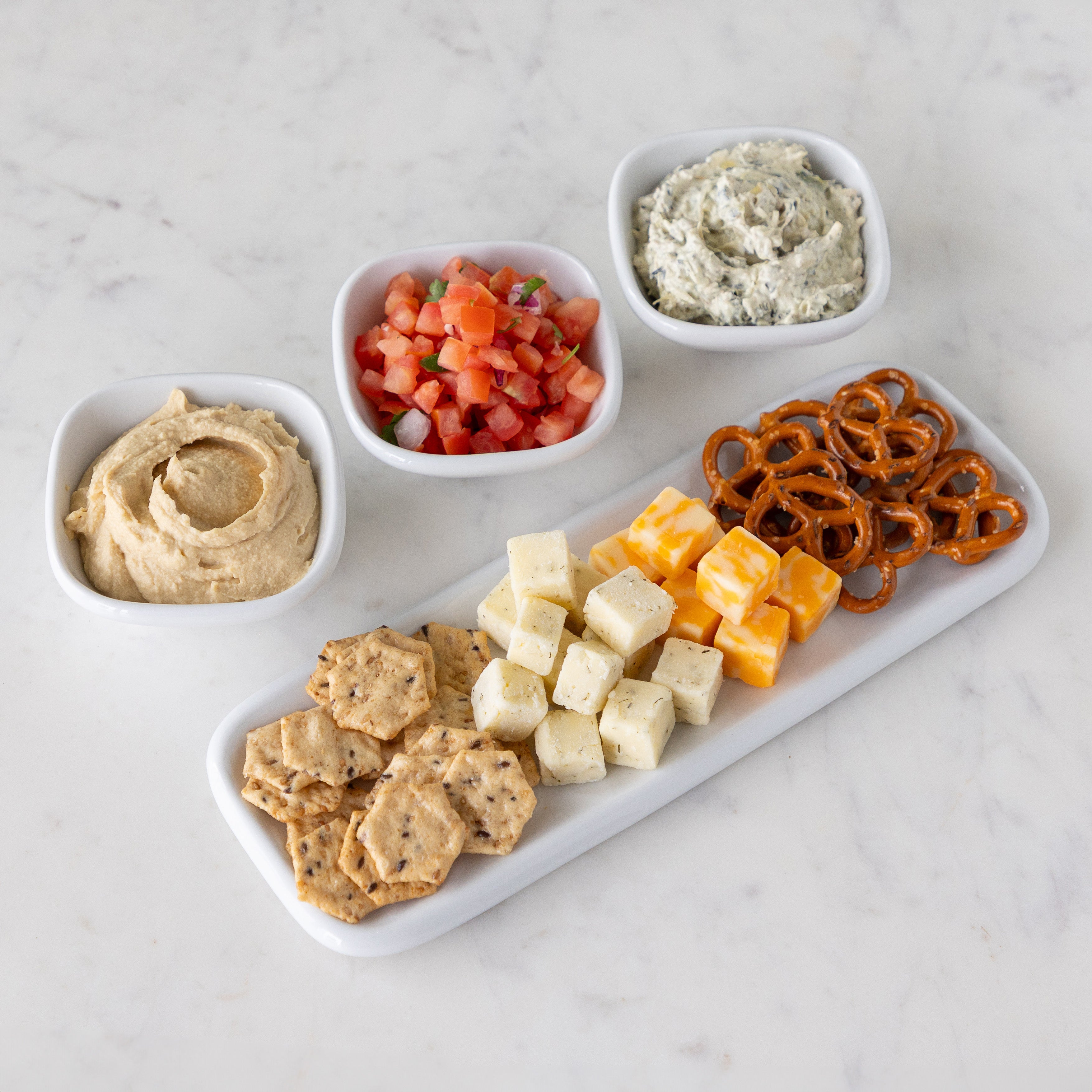Assorted dips in white bowls and assorted snacks including crackers, cheese cubes, and pretzels on a white marble tray.