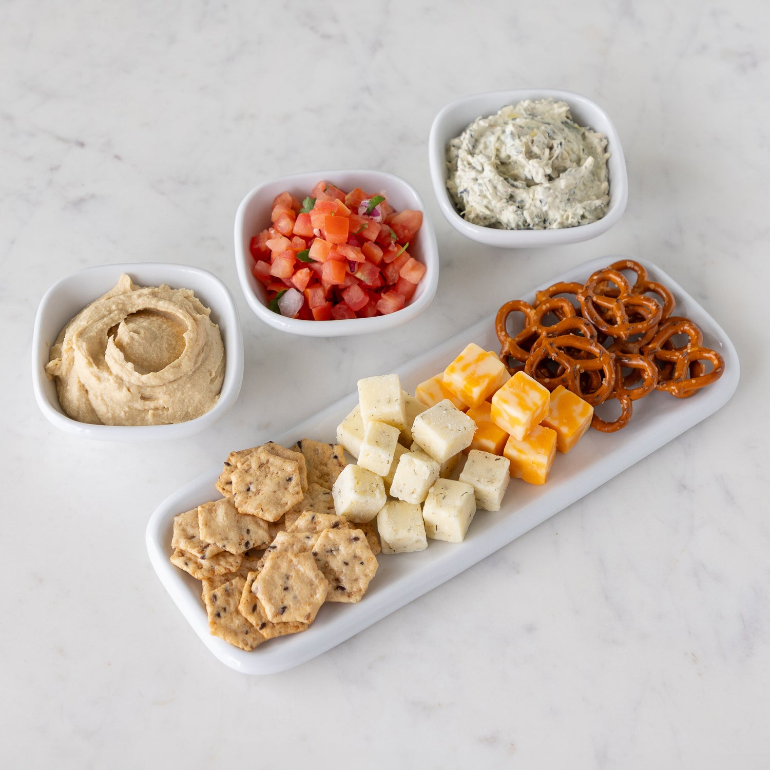 Assorted dips in white bowls and assorted snacks including crackers, cheese cubes, and pretzels on a white marble tray.