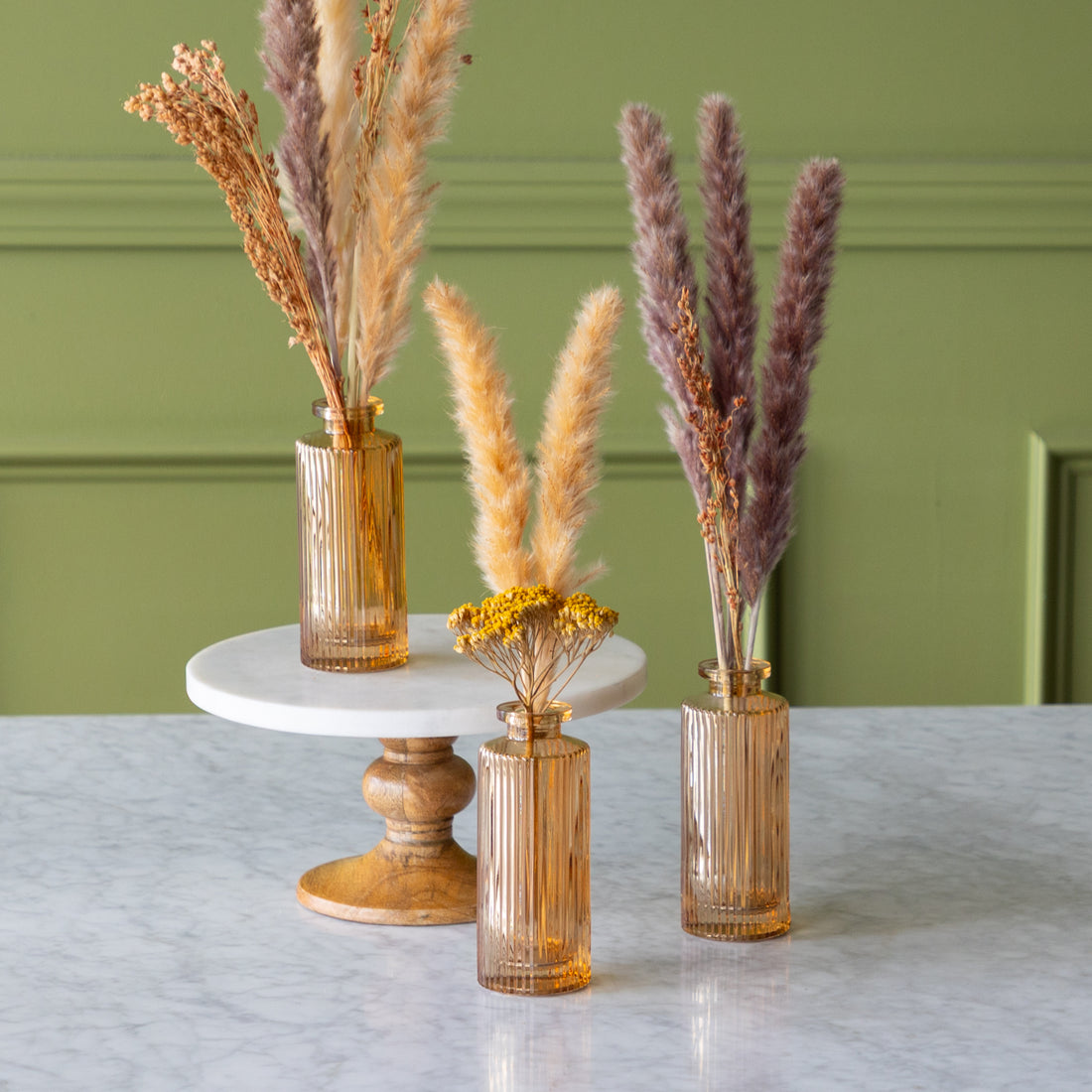 Three Amber Pleated Glass Bottle Bud Vases with dried plants on a marble surface against a green wall.