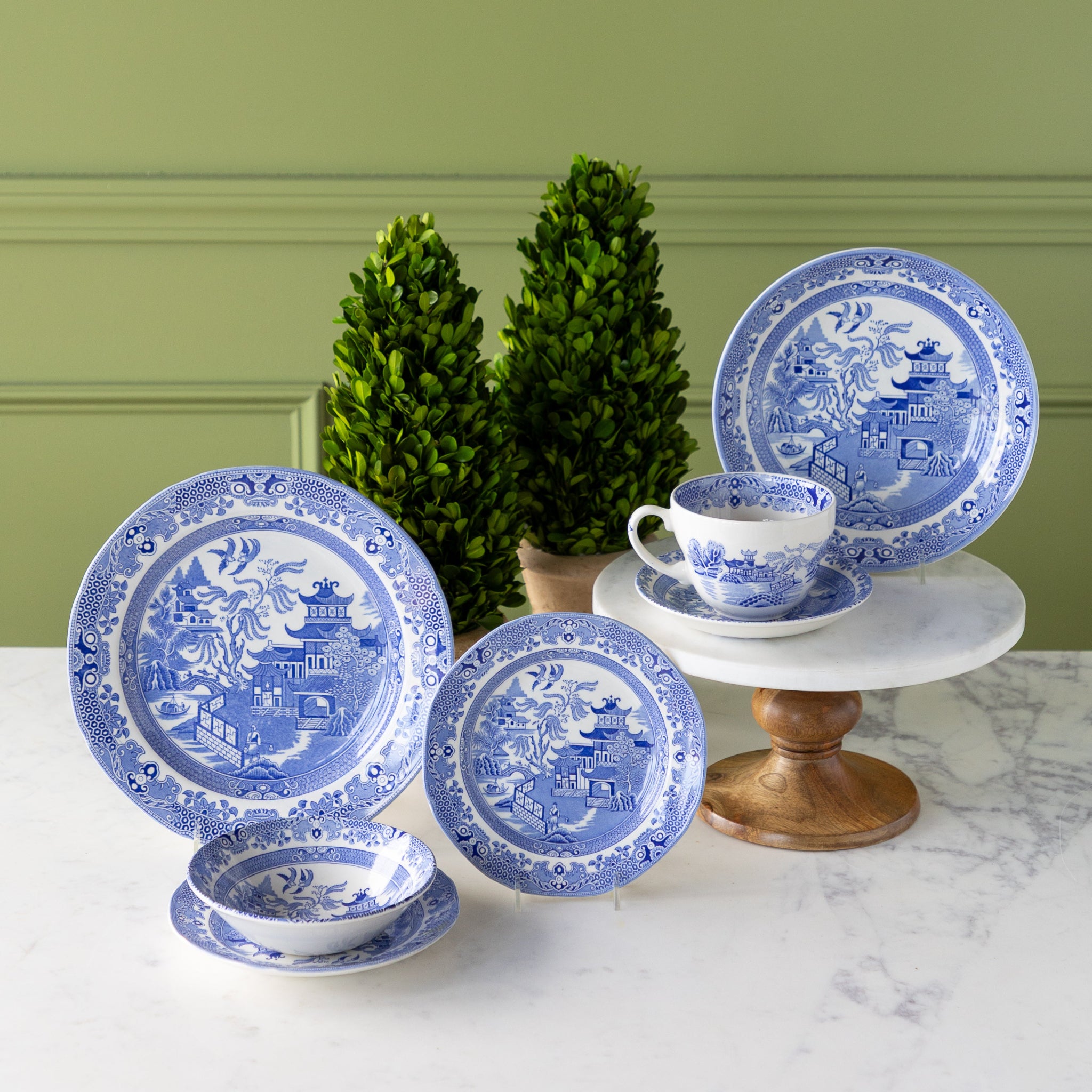 Burleigh Blue Willow Dinnerware displayed on a marble table with topiary plants behind it.